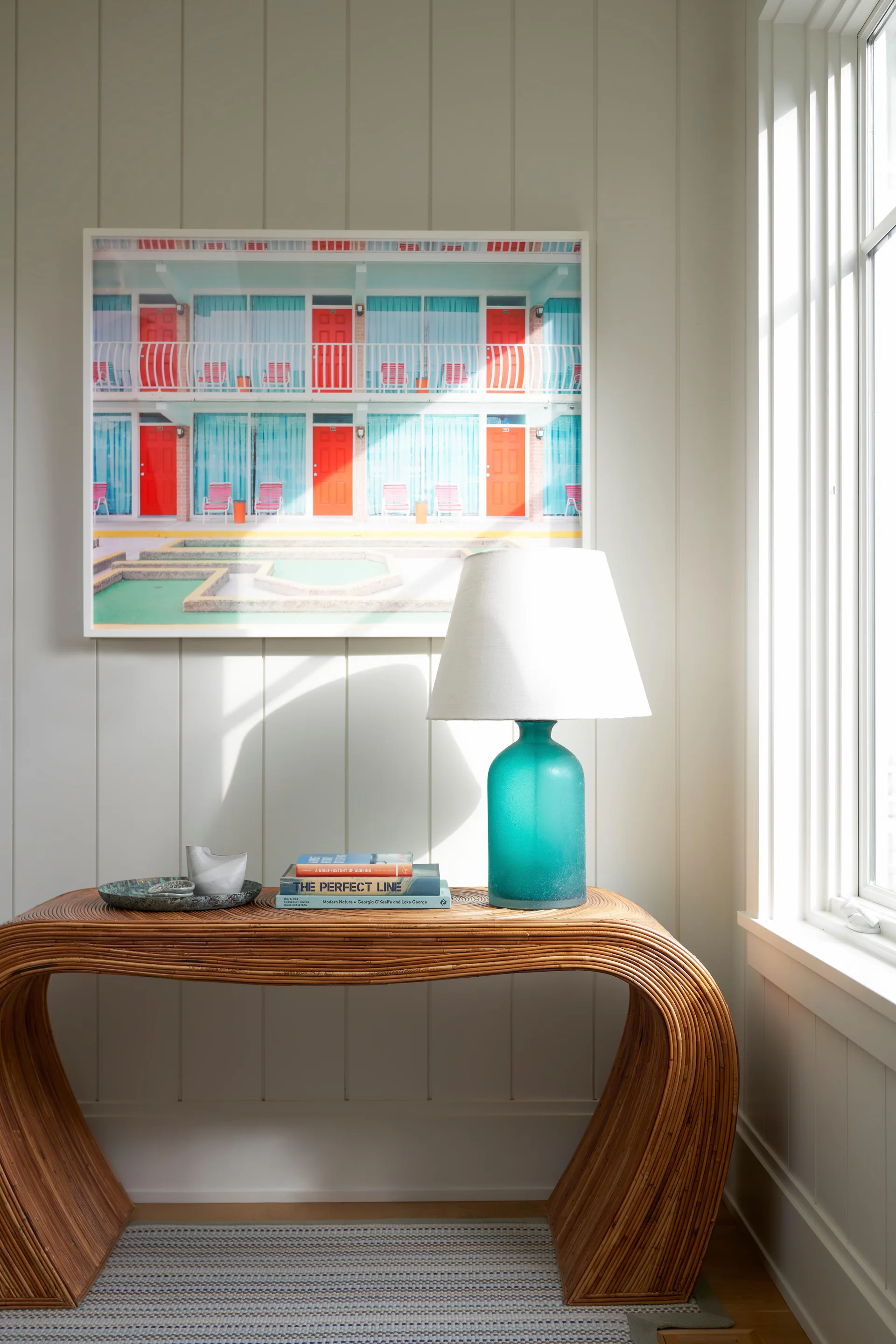 A curved rattan console table sits against vertical wood paneled walls next to a window, holding a green glass lamp, books, and framed beachy artwork.