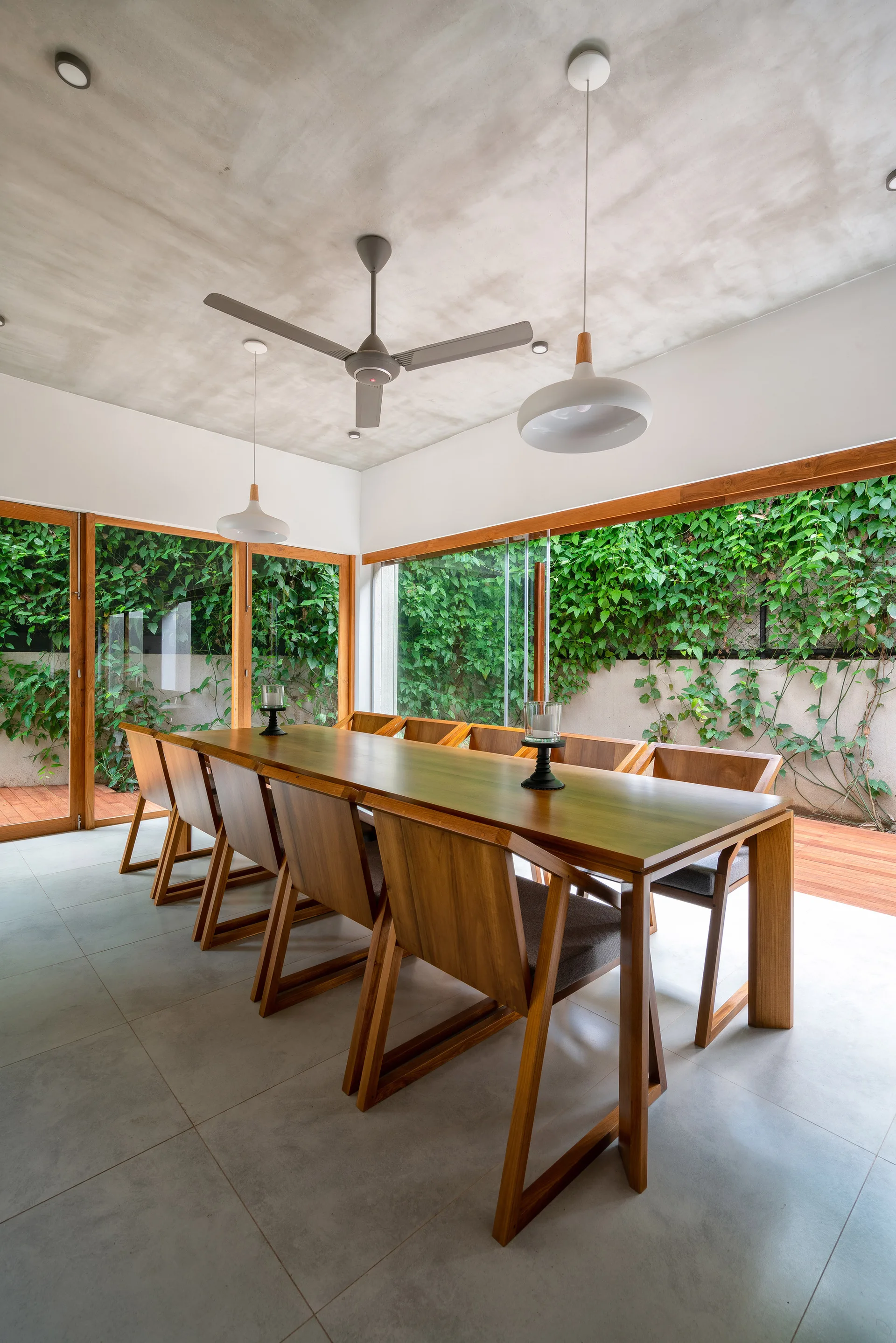 A minimalist dining room featuring a long wooden table and chairs beneath a concrete ceiling, surrounded by glass doors opening to green foliage.