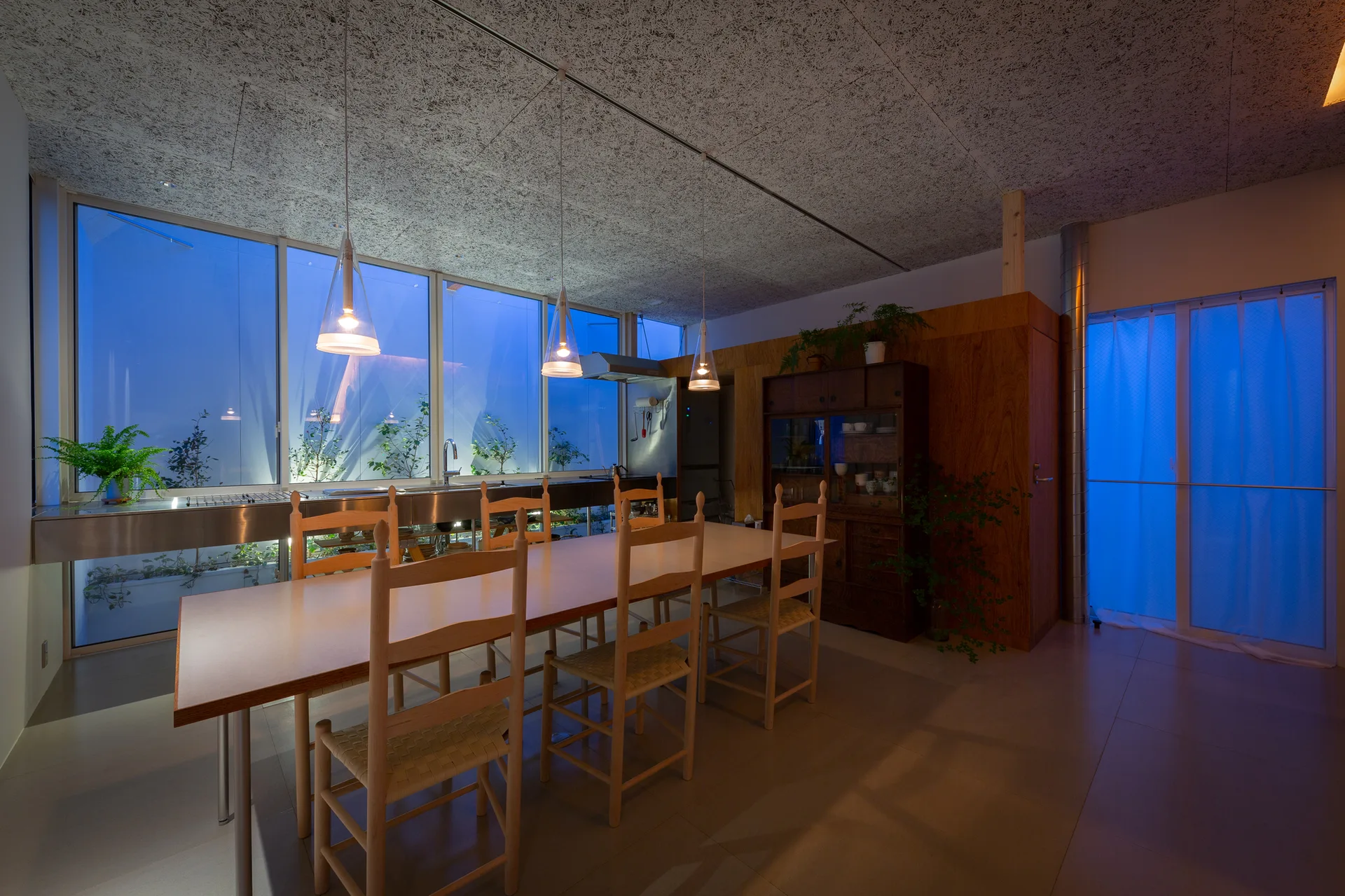 A minimalist dining room featuring a long wooden table with ladderback chairs beneath glass pendant lights and a textured ceiling.
