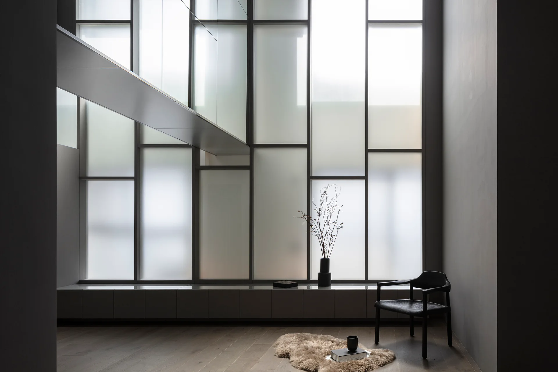 A minimalist grey room features a towering frosted glass grid window wall, a low cabinet, a black chair, and a sheepskin rug on wood floors.