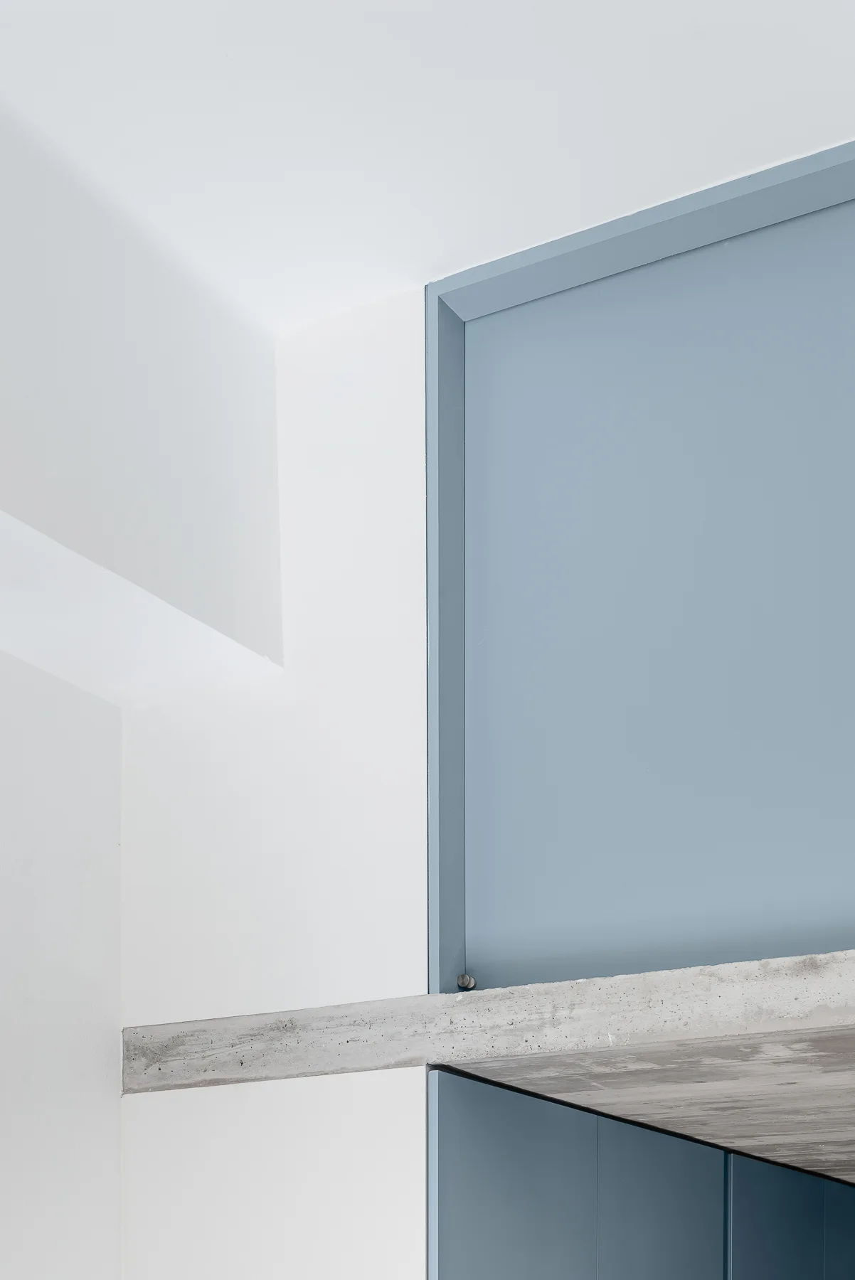 A minimalist interior detail showing a smooth blue panel adjacent to a white wall and a raw exposed concrete beam.