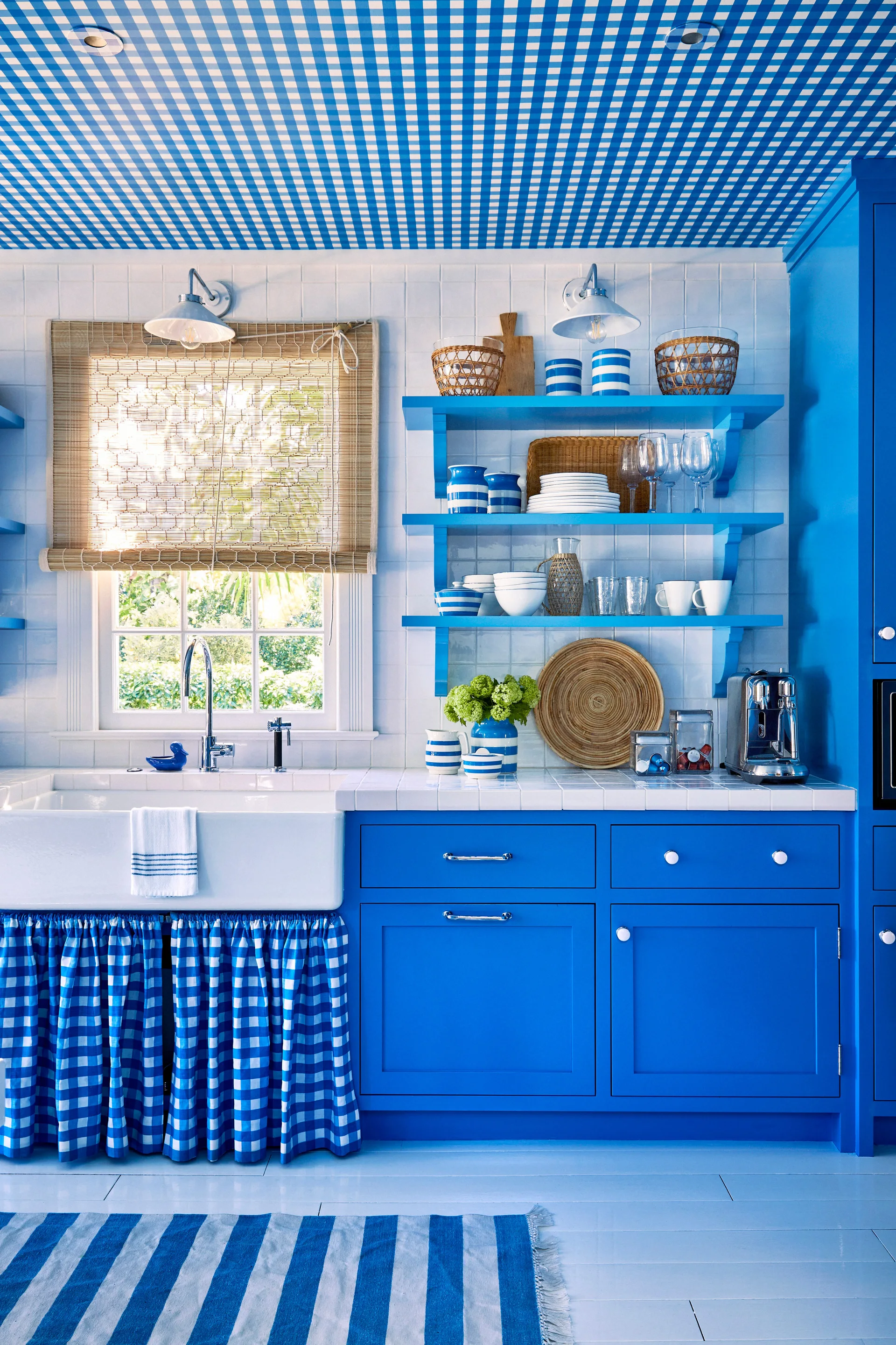 A blue kitchen featuring a gingham-patterned ceiling, open shelves, a white farmhouse sink with a checkered skirt, and a bamboo window shade.