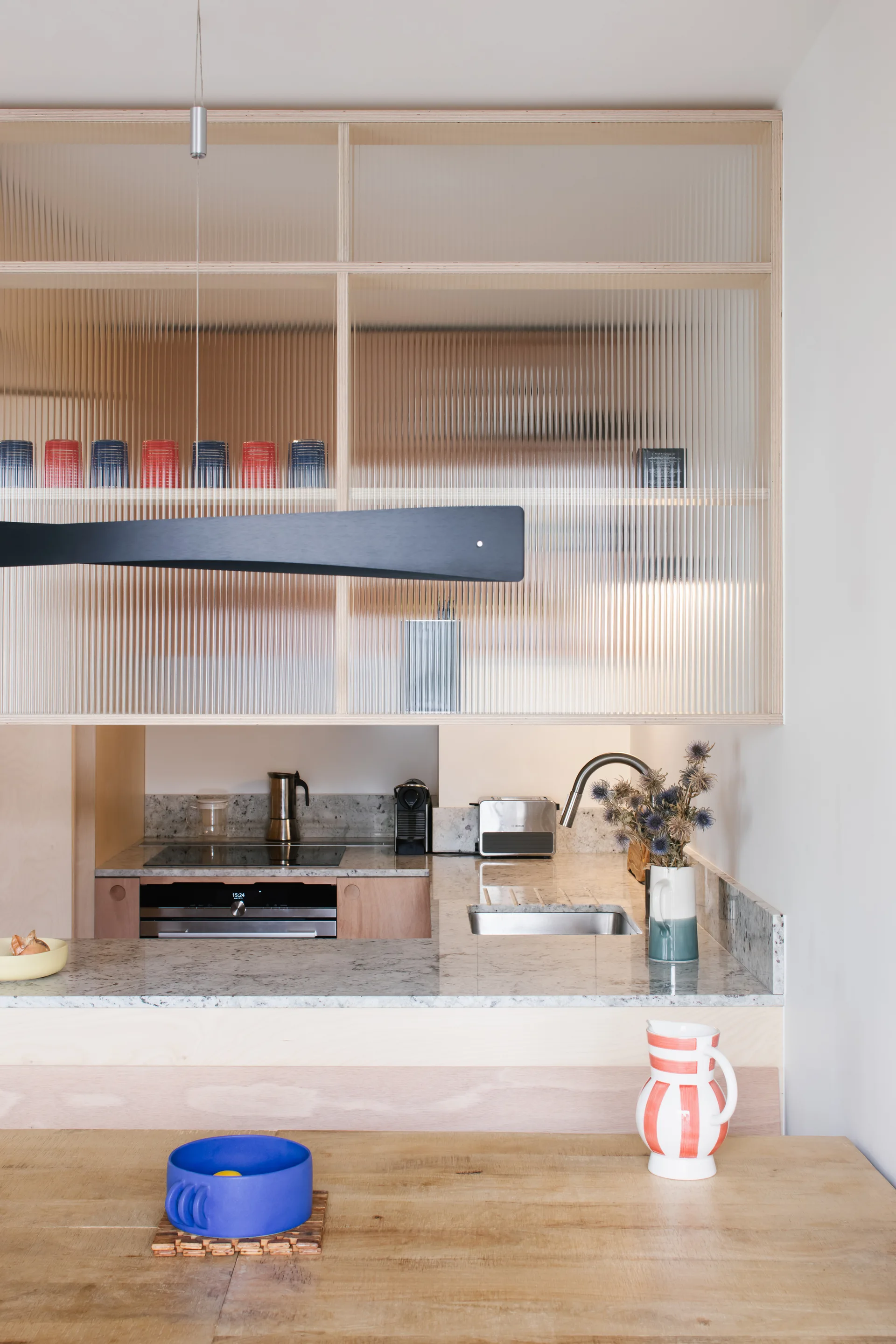 Modern kitchen featuring light wood cabinets with reeded glass doors, a speckled stone countertop with a sink, and a black linear pendant light.