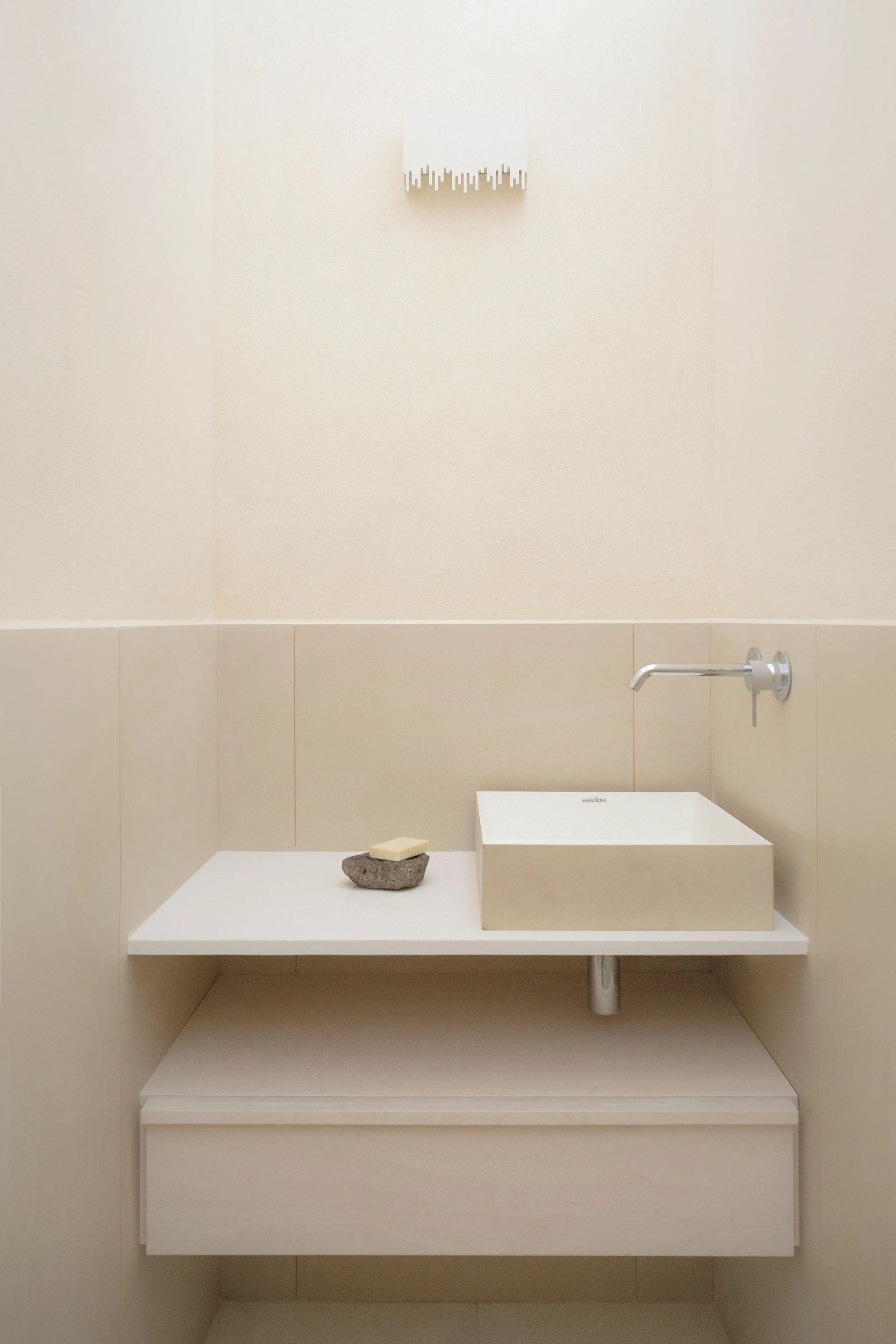 A minimalist beige powder room with a floating console shelf, a white vessel sink, a wall-mounted faucet, and a decorative wall sconce.