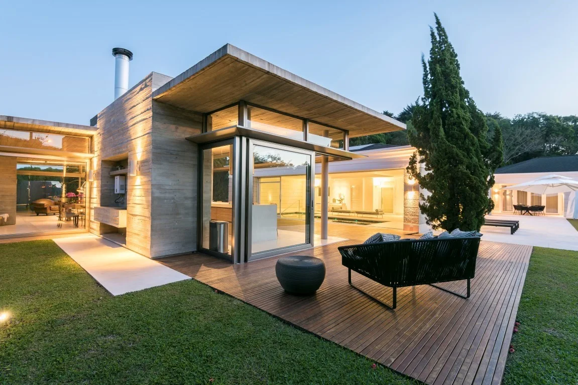 A modern home exterior featuring a wooden deck with a black woven sofa, set against raw concrete walls and large glass doors at dusk.