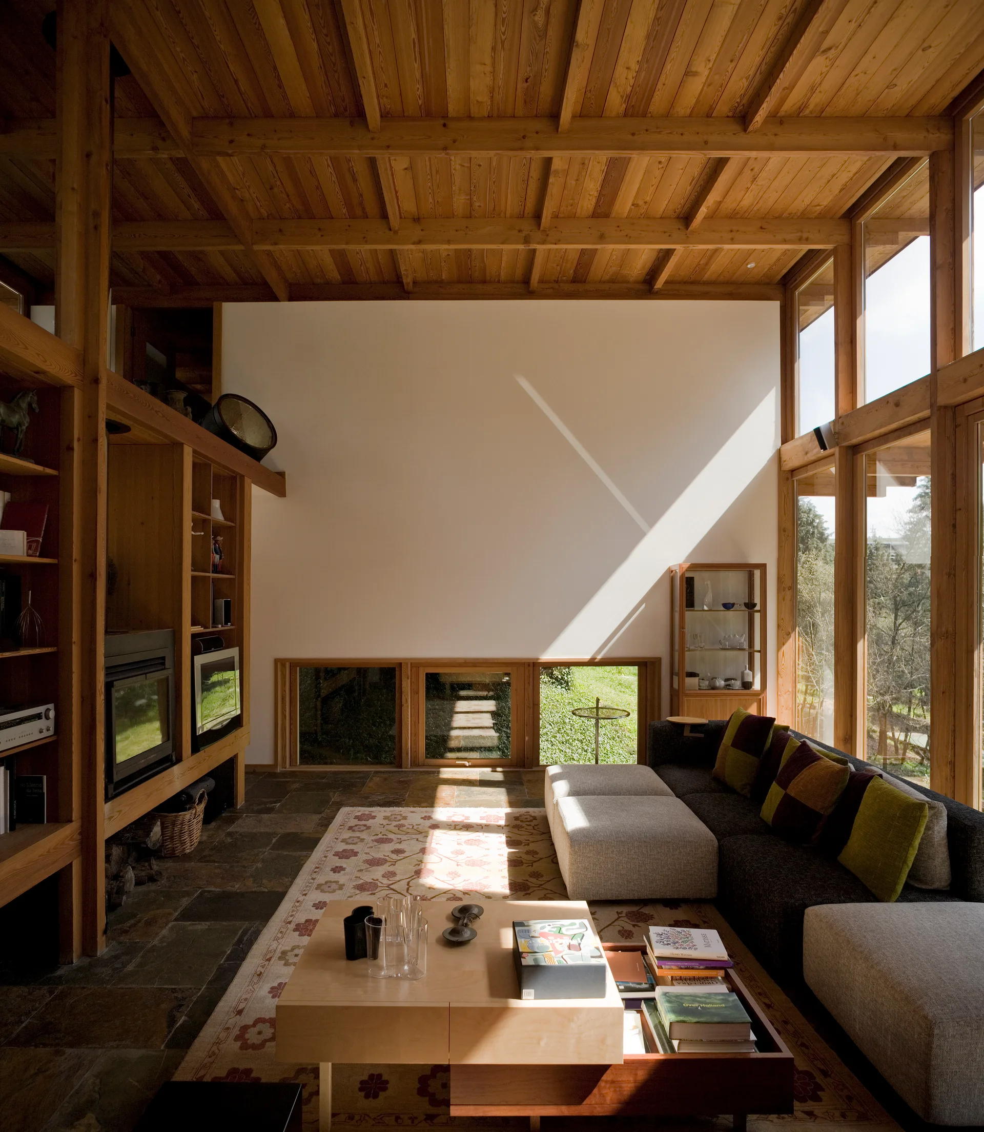 A spacious living room featuring a grid timber ceiling, stone tile flooring, a grey modular sofa, and floor-to-ceiling windows.