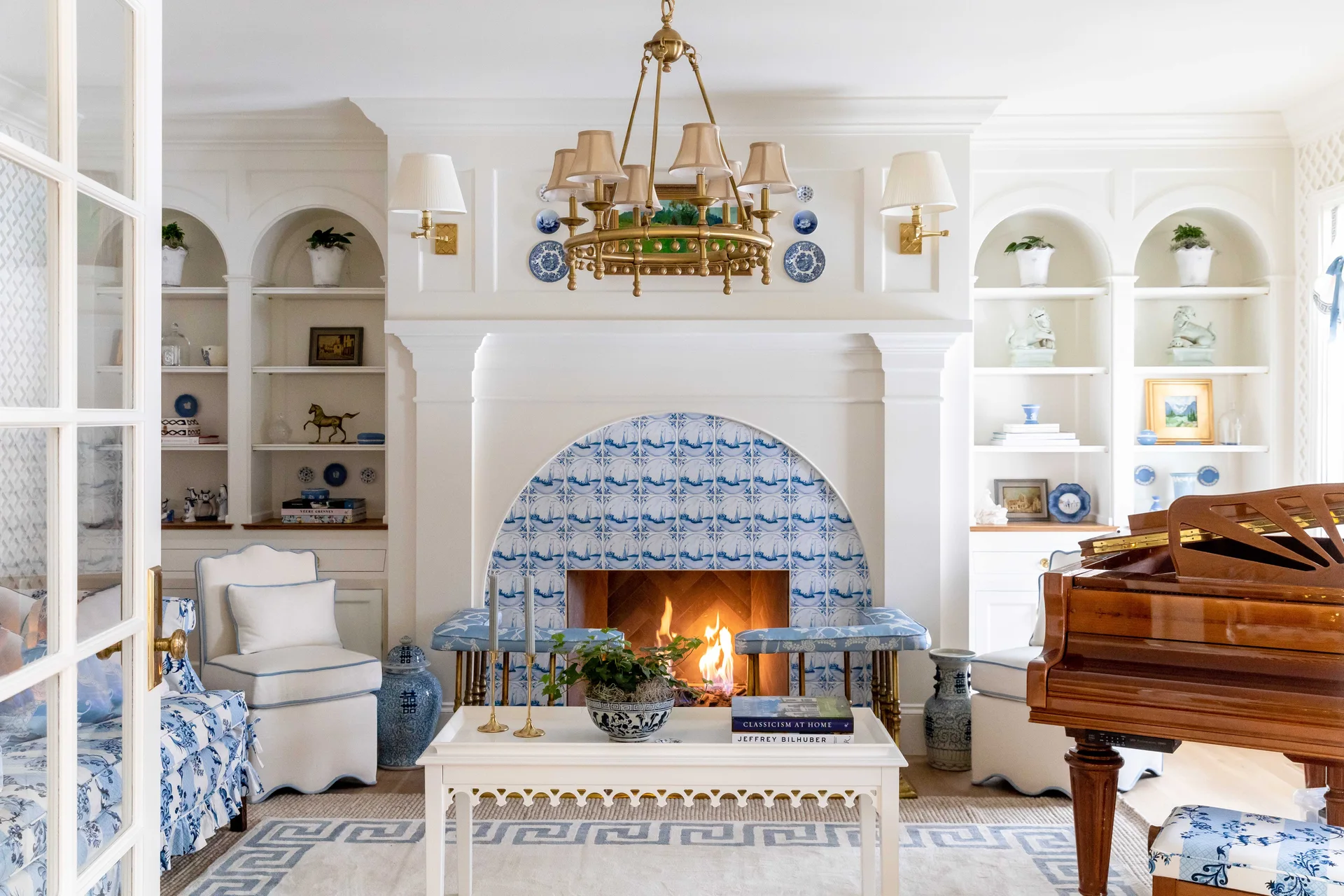 A white living room features an arched fireplace with blue and white tiles, built-in bookshelves, and a classic wood grand piano.
