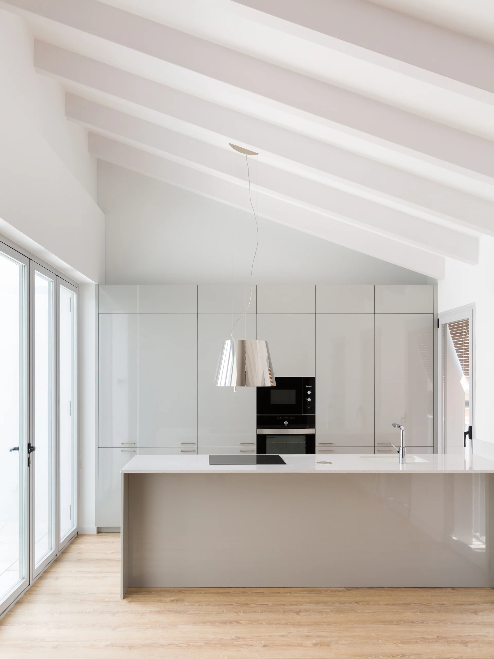 A minimalist kitchen features glossy light-gray cabinets, a central island with a sink and cooktop, and a vaulted ceiling with painted white beams.