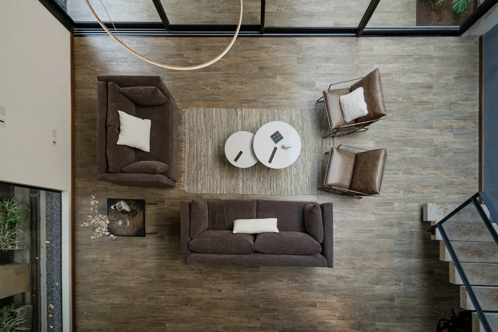 A high-angle aerial view of a living room featuring two brown sofas, two leather armchairs, and two round white nested coffee tables on a woven rug.