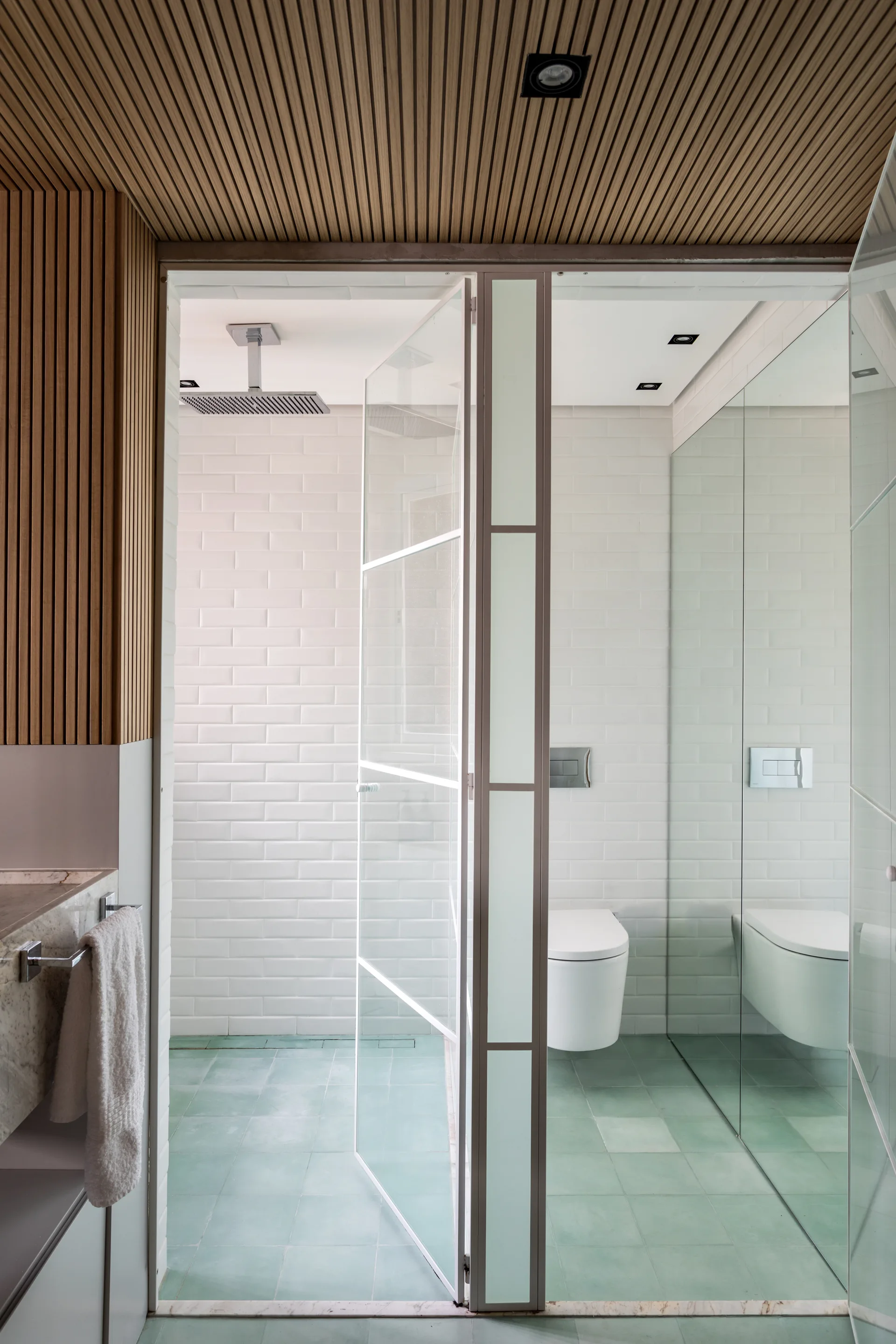 A minimalist bathroom features a linear wood slat ceiling, white subway tile walls, and a glass-enclosed shower with light green floor tiles.