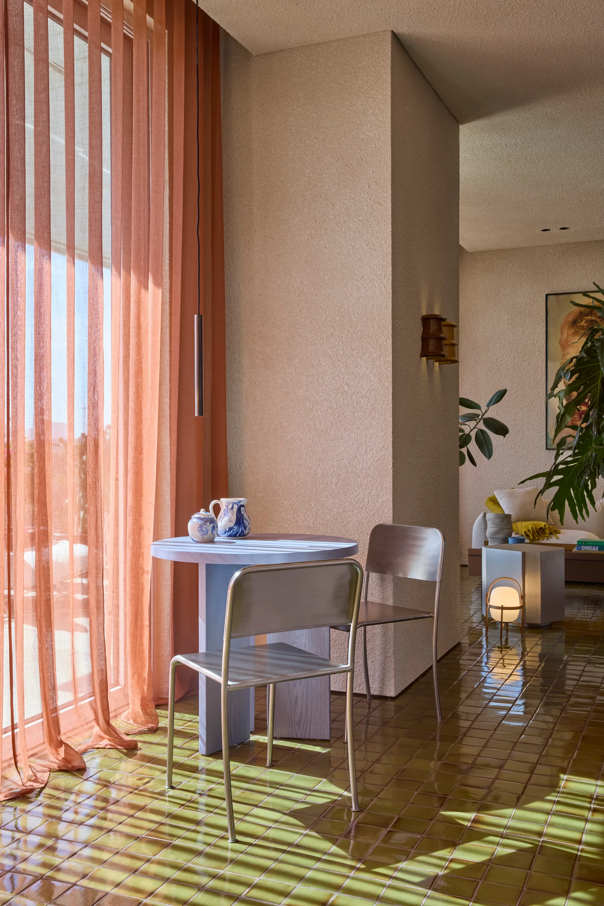 A small dining table with metal chairs sits on glossy olive green floor tiles next to windows with long sheer terracotta curtains.