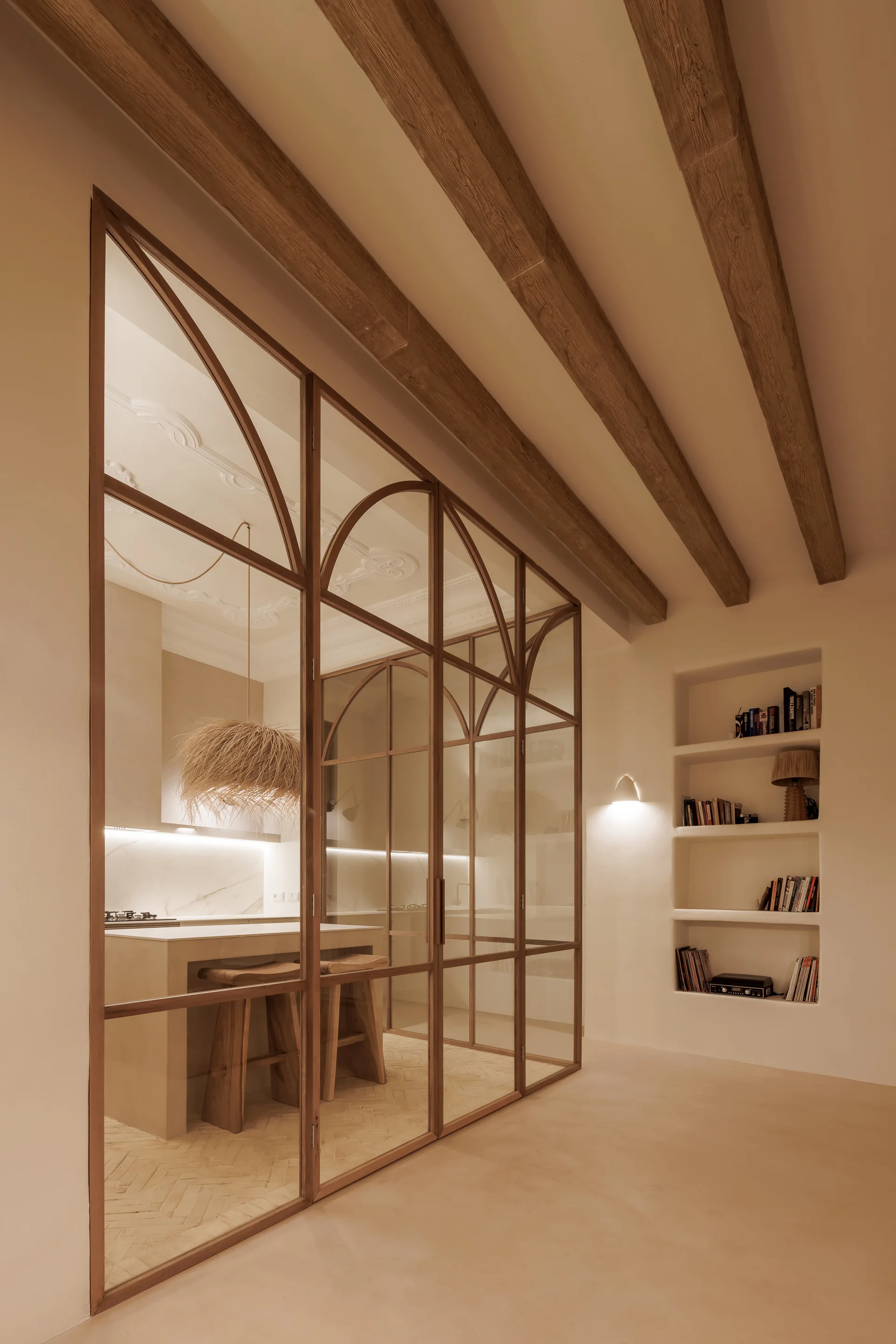 A view of a modern kitchen through a large arched glass partition with exposed wooden ceiling beams and built-in shelving.