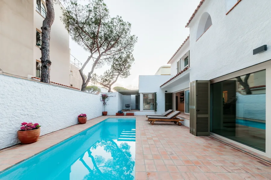 A long blue swimming pool sits in a Mediterranean-style courtyard with terracotta tile flooring and a textured white stucco exterior wall.