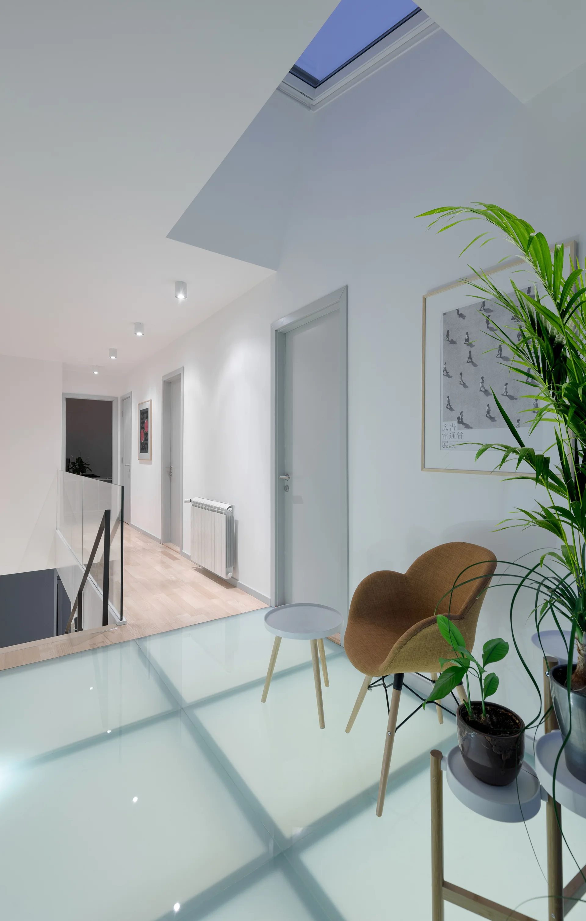 A modern upstairs landing features a translucent glass floor, a brown armchair next to a plant, and grey doors along a white hallway.