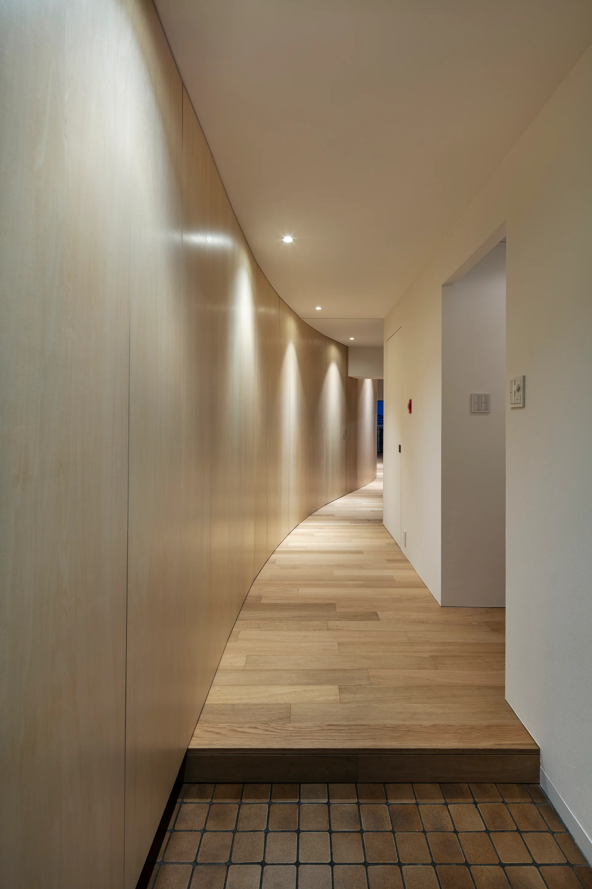 A modern hallway with a long curved wood panel wall illuminated by spotlights, transition step, and tiled flooring.