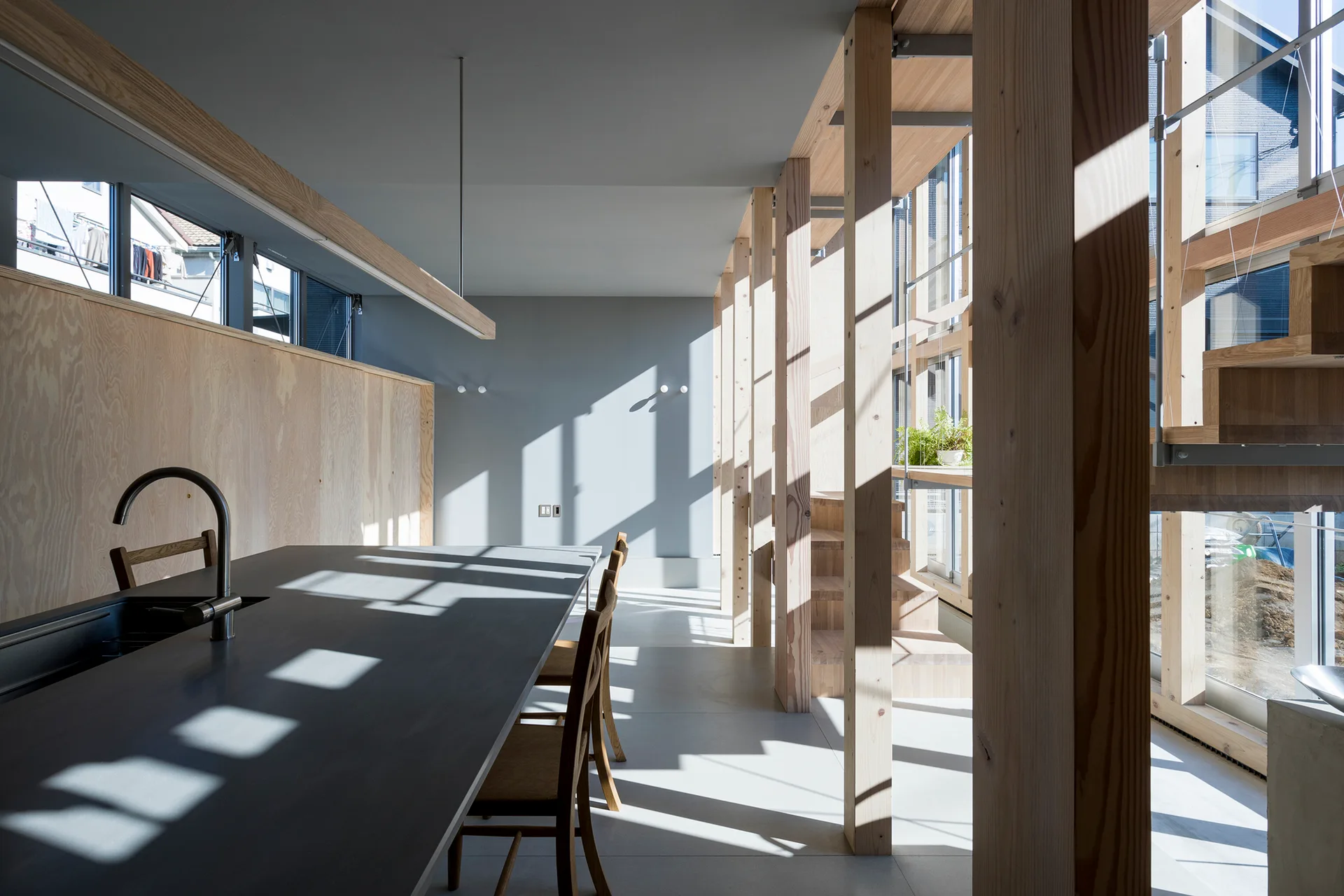 A minimalist kitchen features a dark charcoal island and wooden chairs flanked by exposed structural timber posts and plywood wall panels.