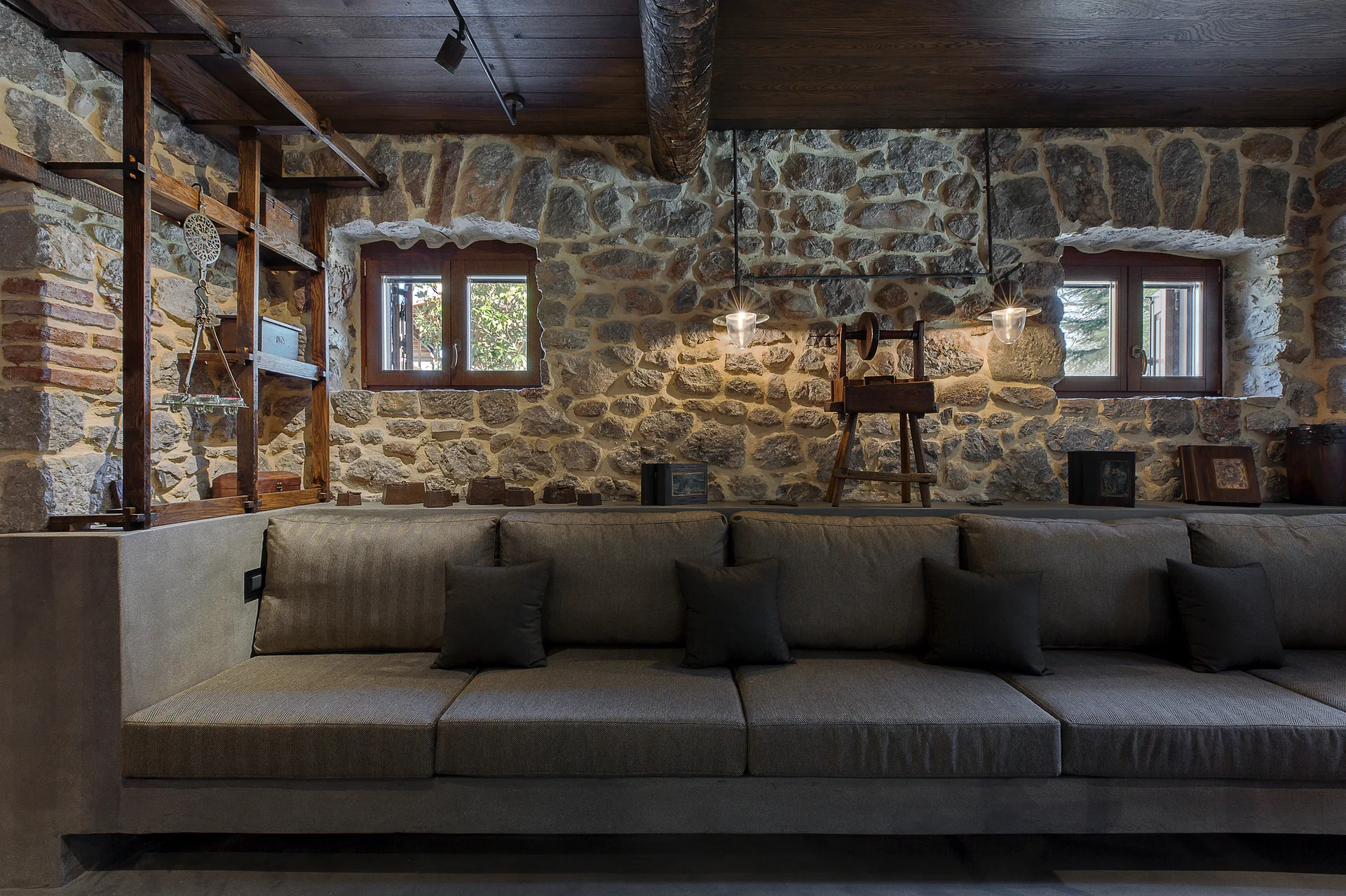A long grey built-in sofa sits against a textured stone wall beneath a dark wood plank ceiling with two small windows and pendant lights.