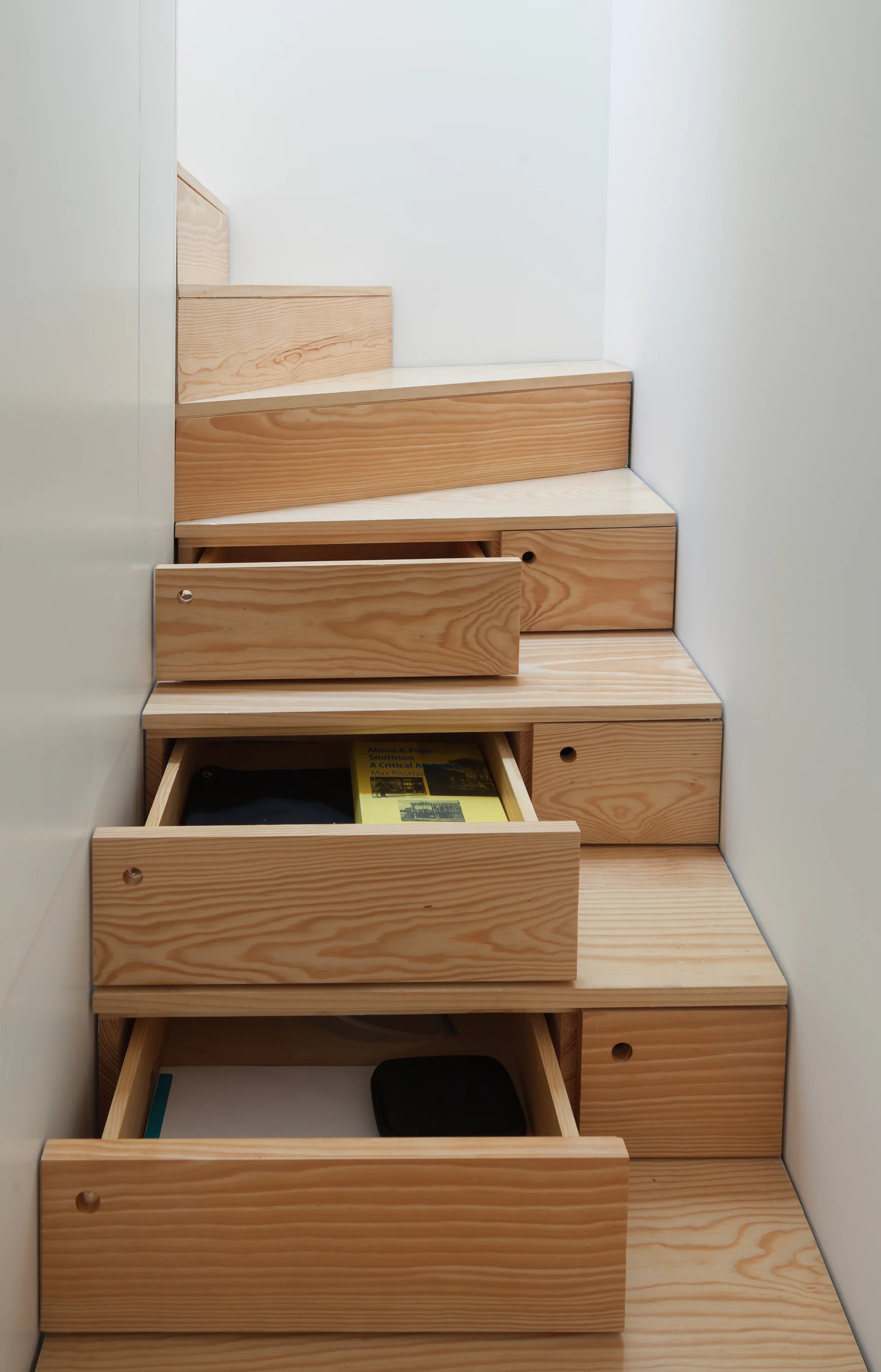 A light wood staircase features built-in storage drawers pulled open to reveal items inside, set between smooth white walls.