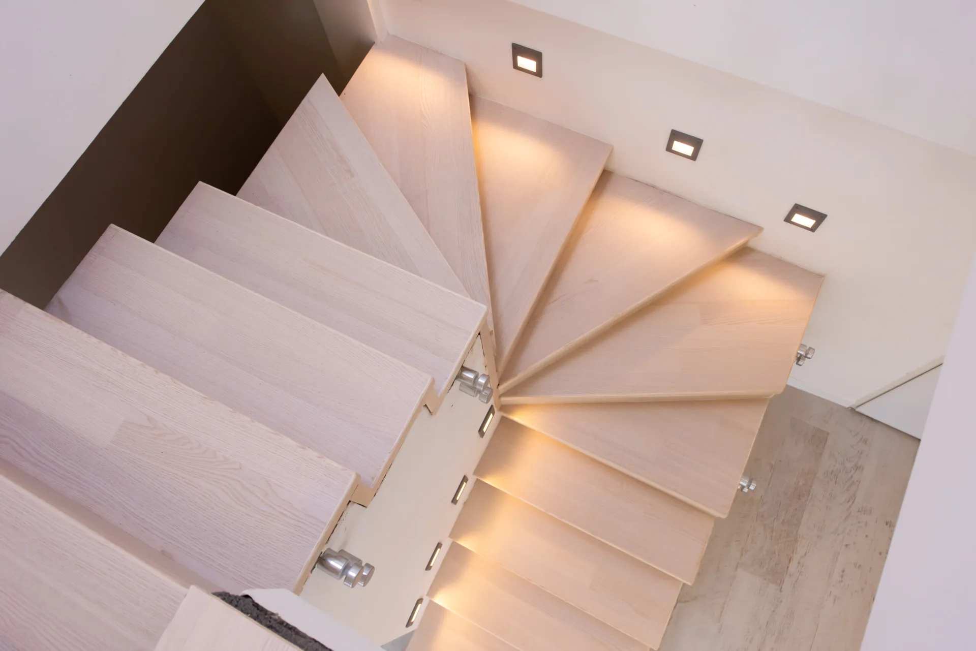 Looking down at a winding staircase with pale wood treads and recessed square lights illuminating the steps.