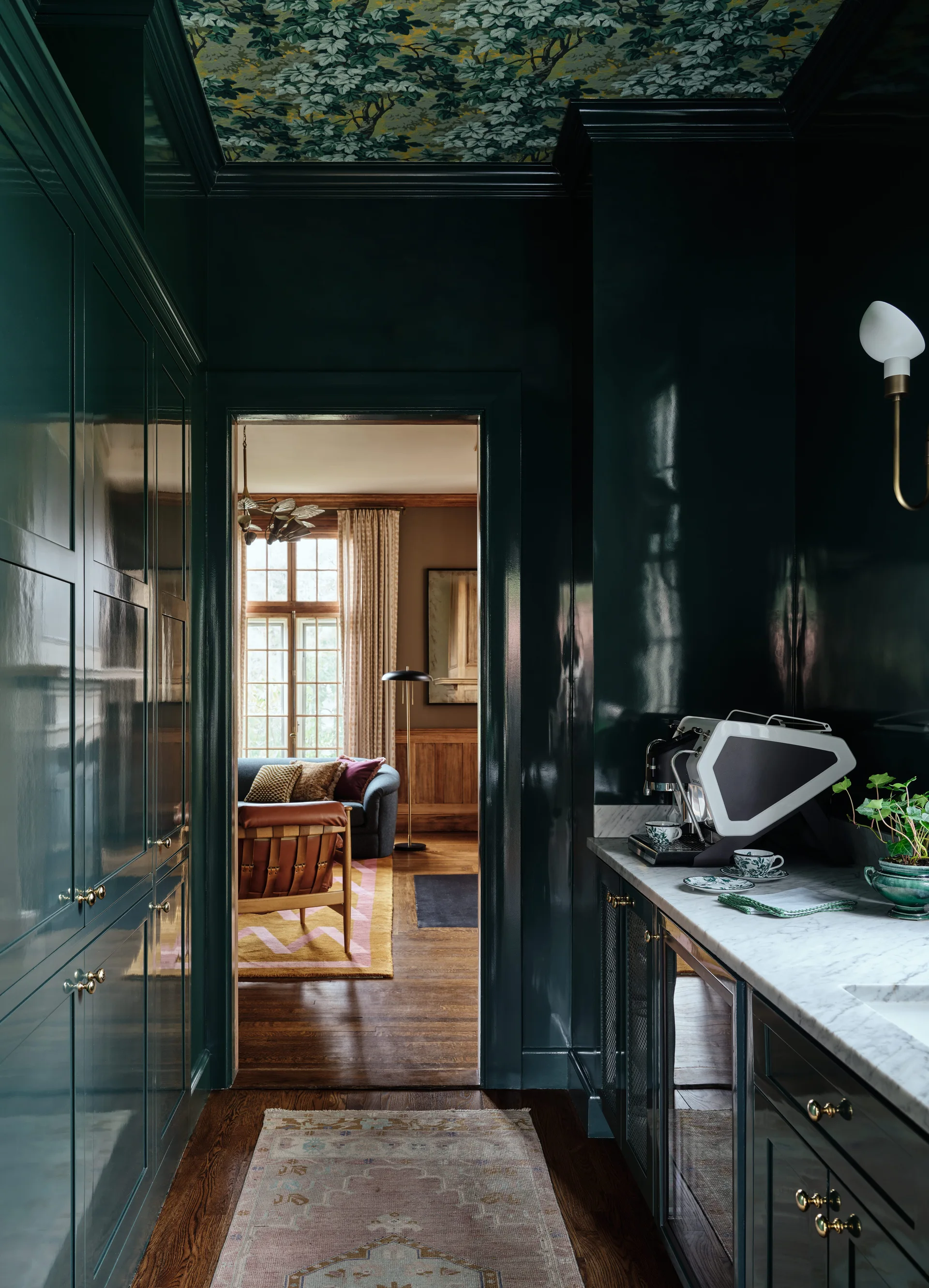 High-gloss dark green kitchen pantry with a white marble countertop, leafy ceiling wallpaper, and a view into a living room.