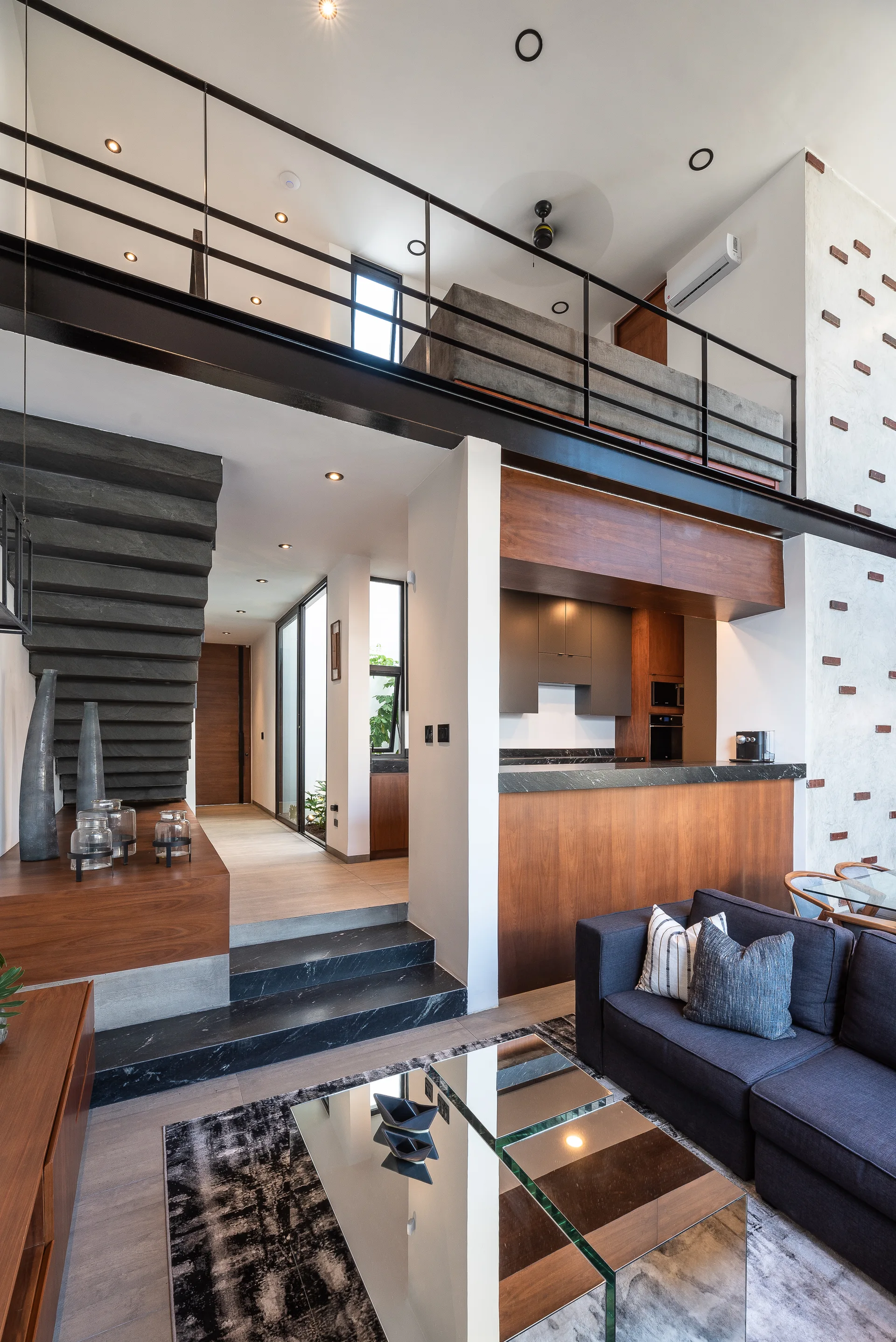 A modern loft living room features a dark gray sofa, a mirrored coffee table, and a black stone staircase leading up to a mezzanine level.