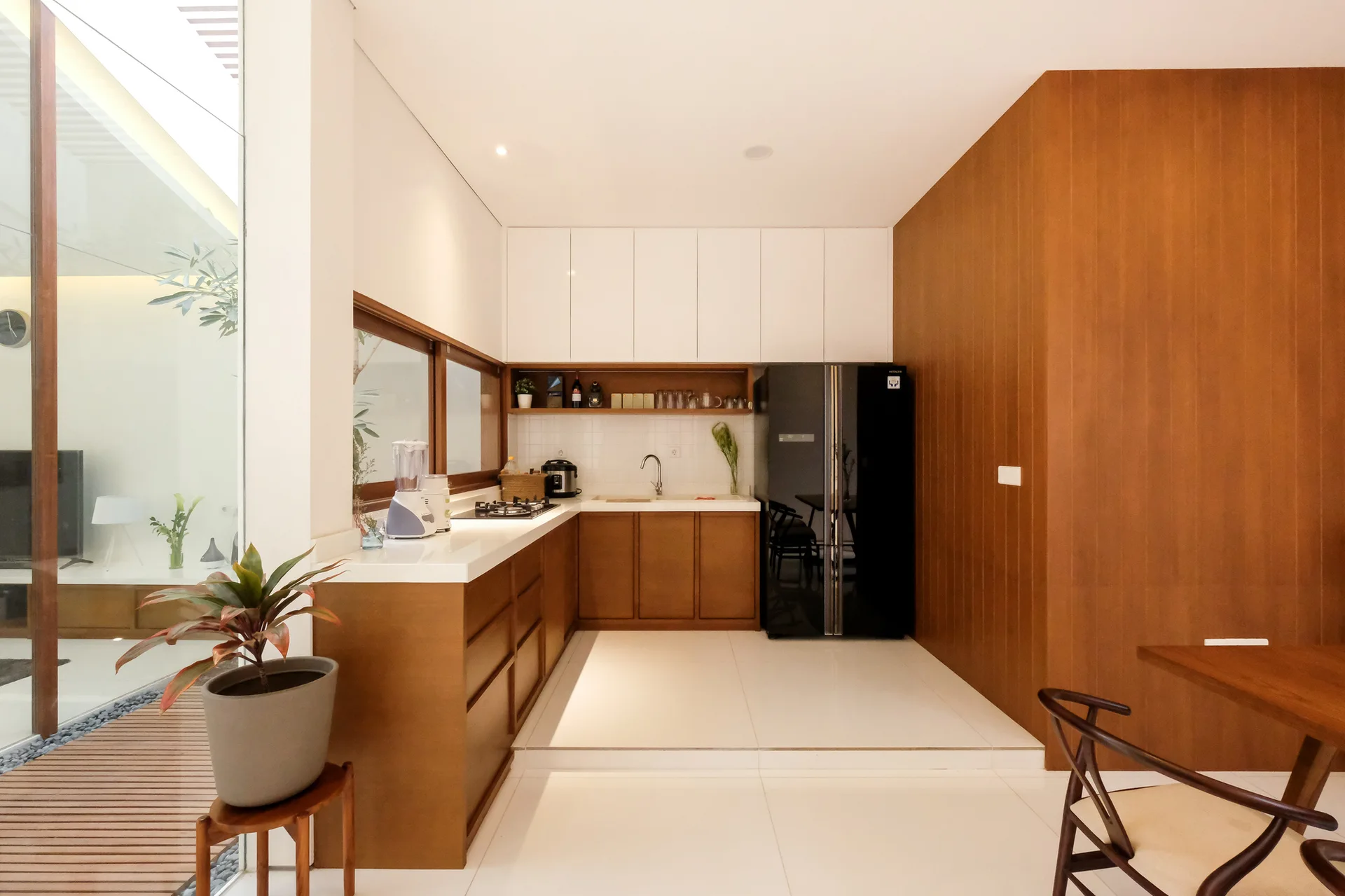 A modern L-shaped kitchen features wood-toned lower cabinets, flat white upper cabinets, a black refrigerator, and a window overlooking an outdoor patio.