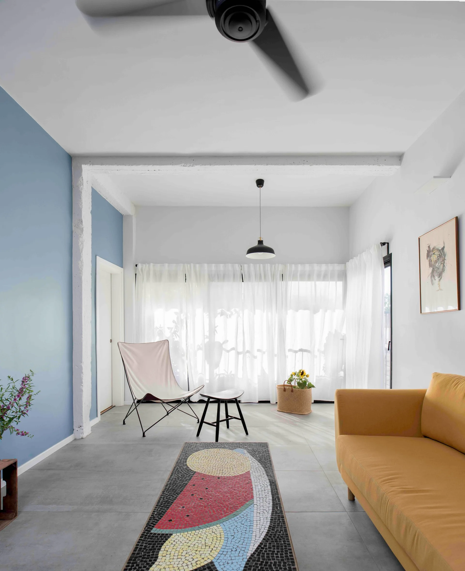 A bright living room features a yellow fabric sofa, a white butterfly chair, a mosaic coffee table, and a periwinkle blue accent wall.