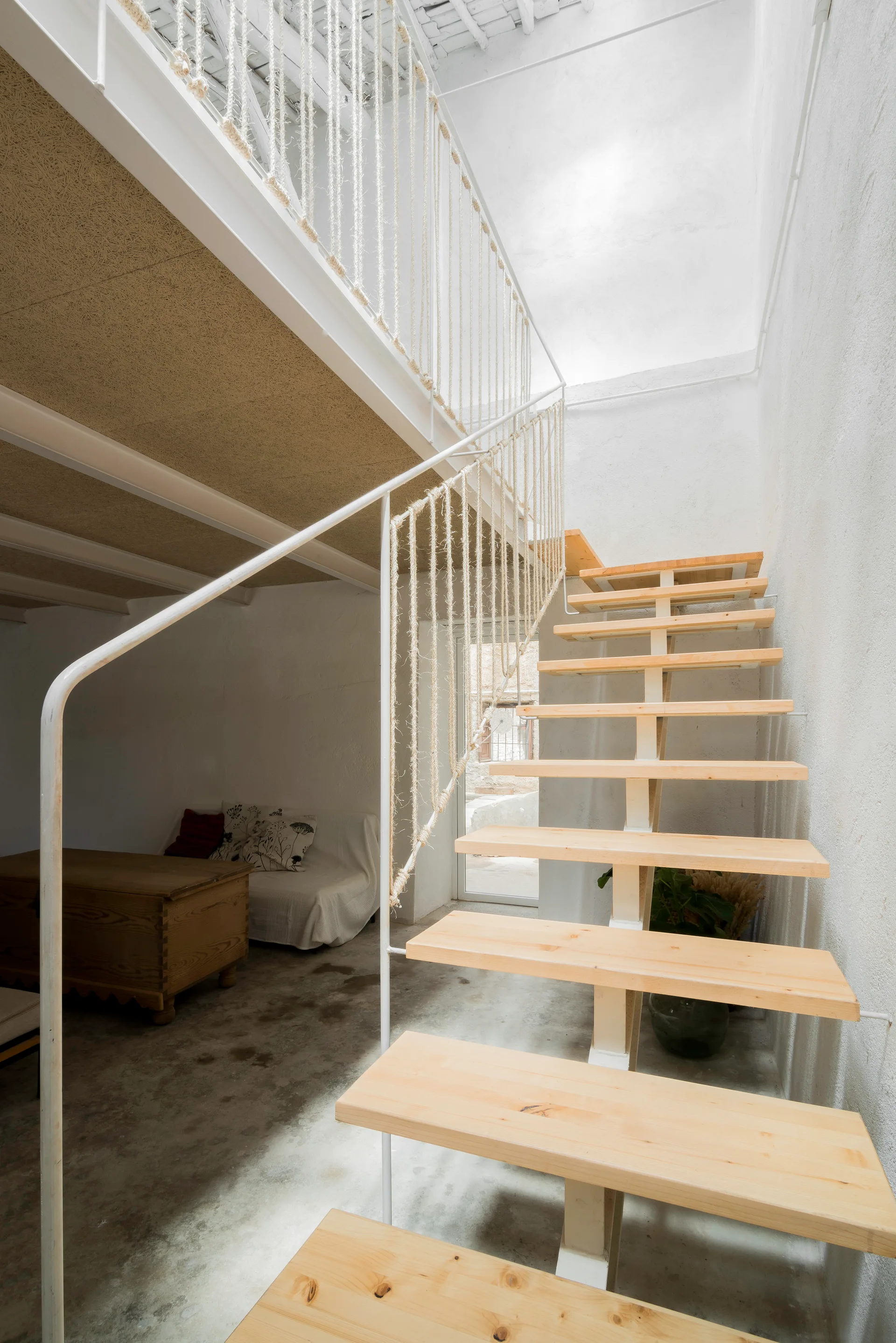 An open-riser wooden staircase with a white metal and rope railing stands against a textured white wall in a minimalist loft.