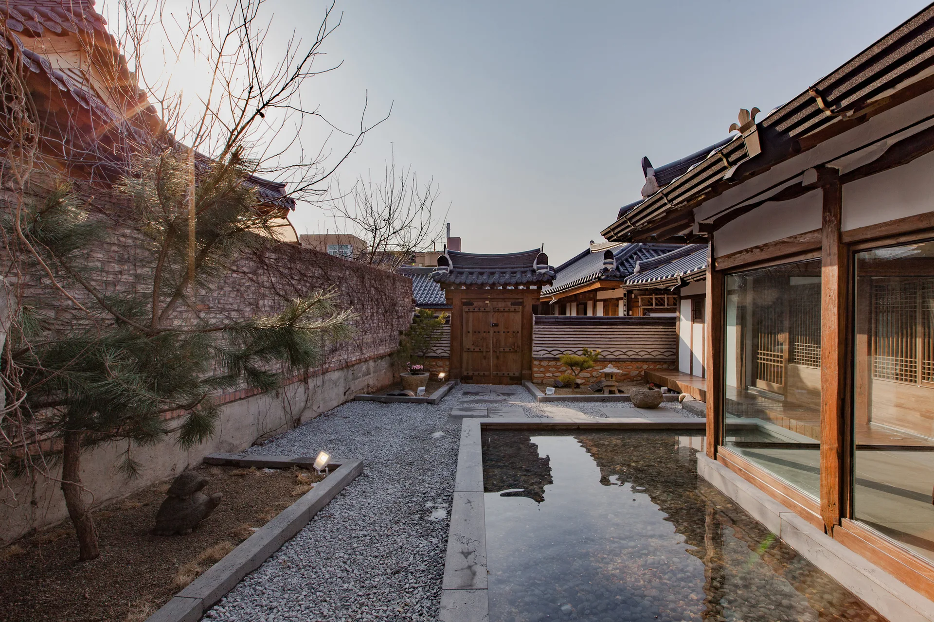 A traditional courtyard features a long reflecting pool, grey gravel ground, a wooden gate, and a hanok-style building with a tiled roof.