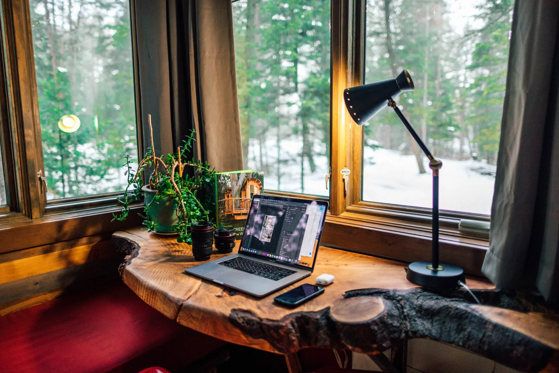 A rustic live-edge wood desk sits in front of a large window overlooking a snowy pine forest, featuring a laptop, black task lamp, and small potted plant.