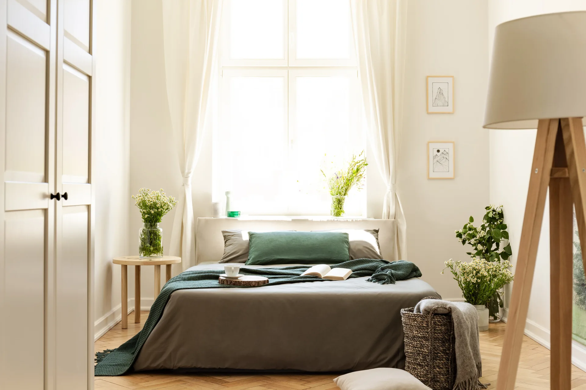 A sunlit bedroom features a bed with forest green bedding, a wooden side table with wildflowers, and a large window with sheer white curtains.