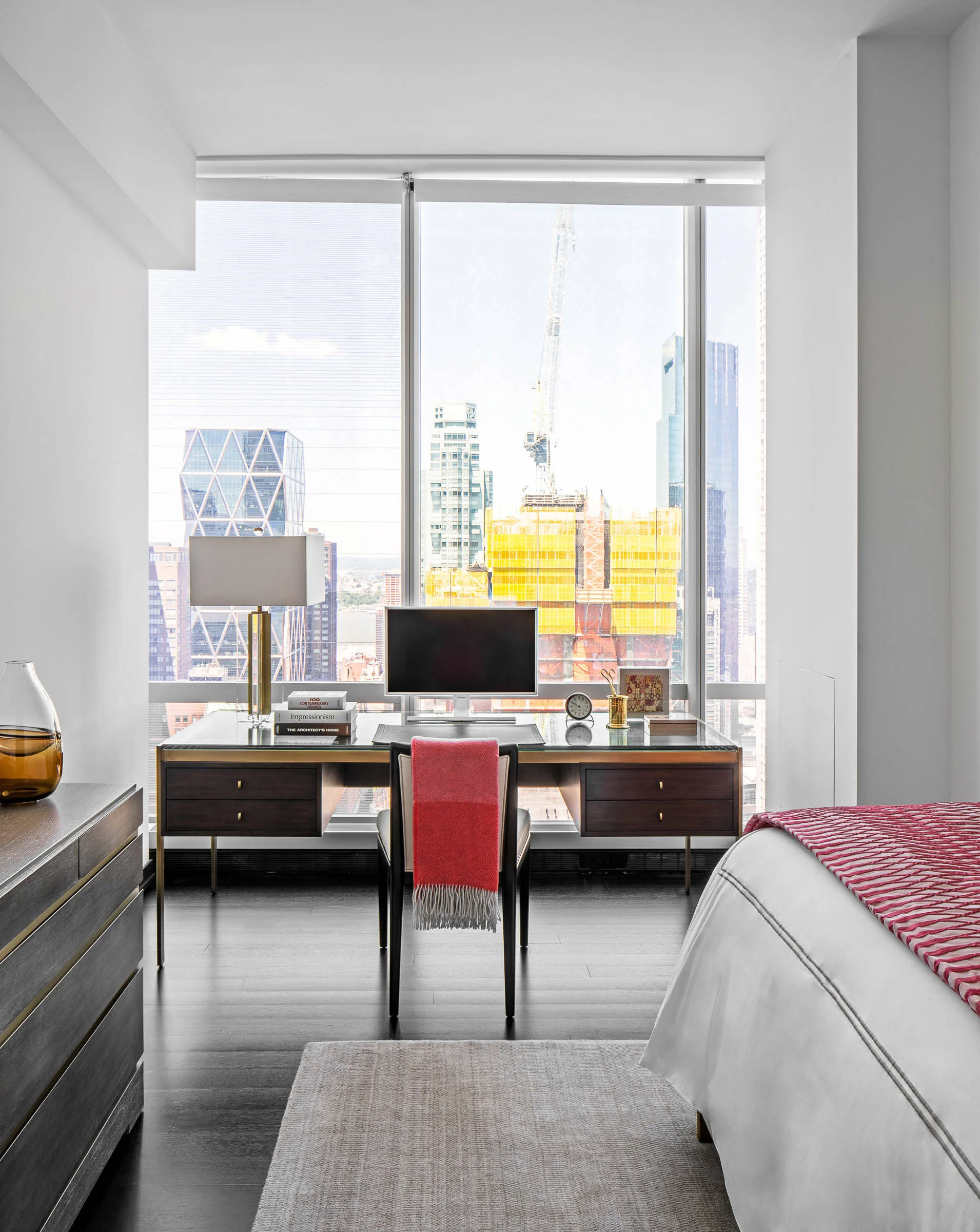 A modern bedroom workspace features a dark wood desk with a glass top positioned in front of a large window overlooking the city.