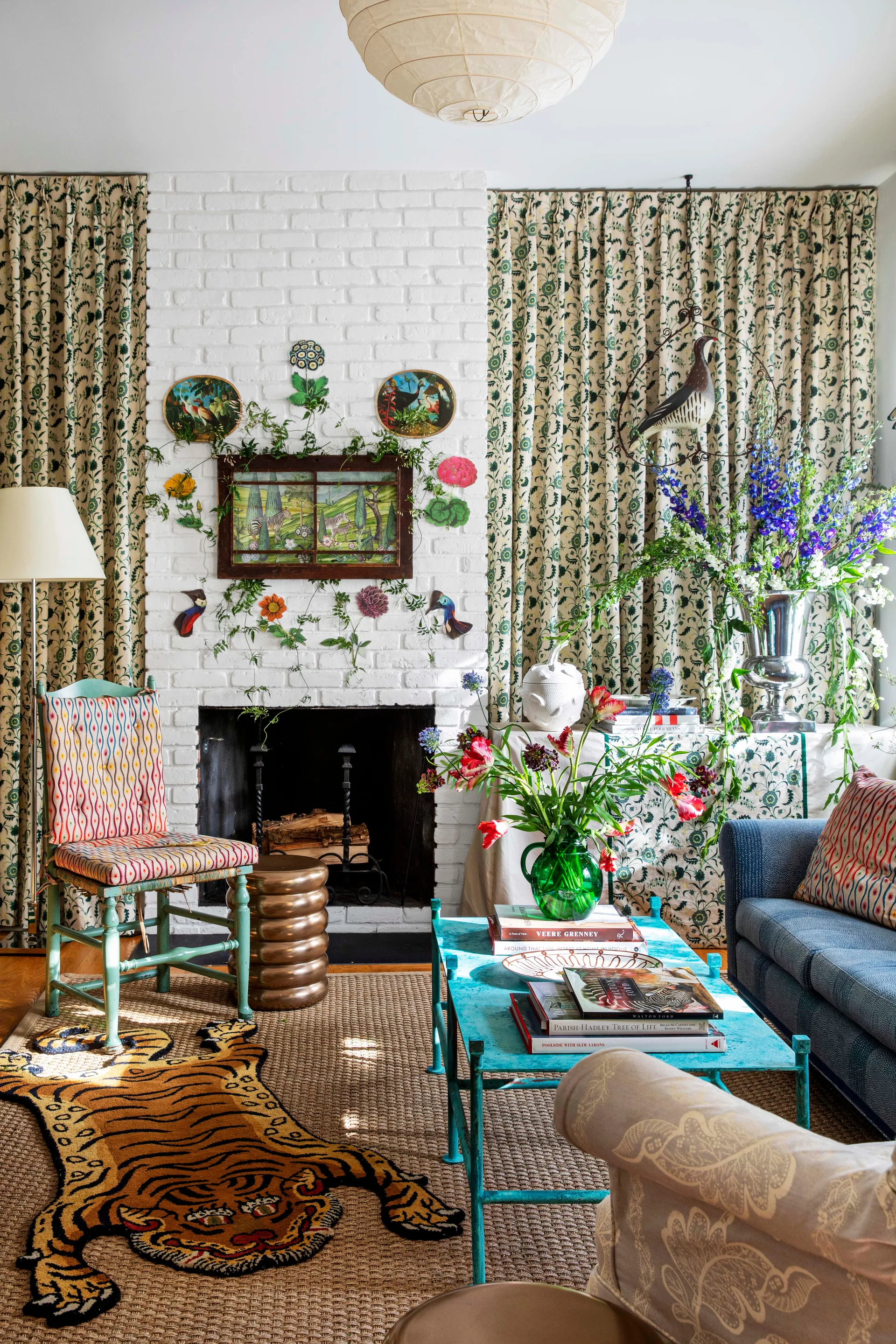 An eclectic living room featuring a white brick fireplace flanked by botanical drapes, a blue coffee table, and a tiger-shaped rug.