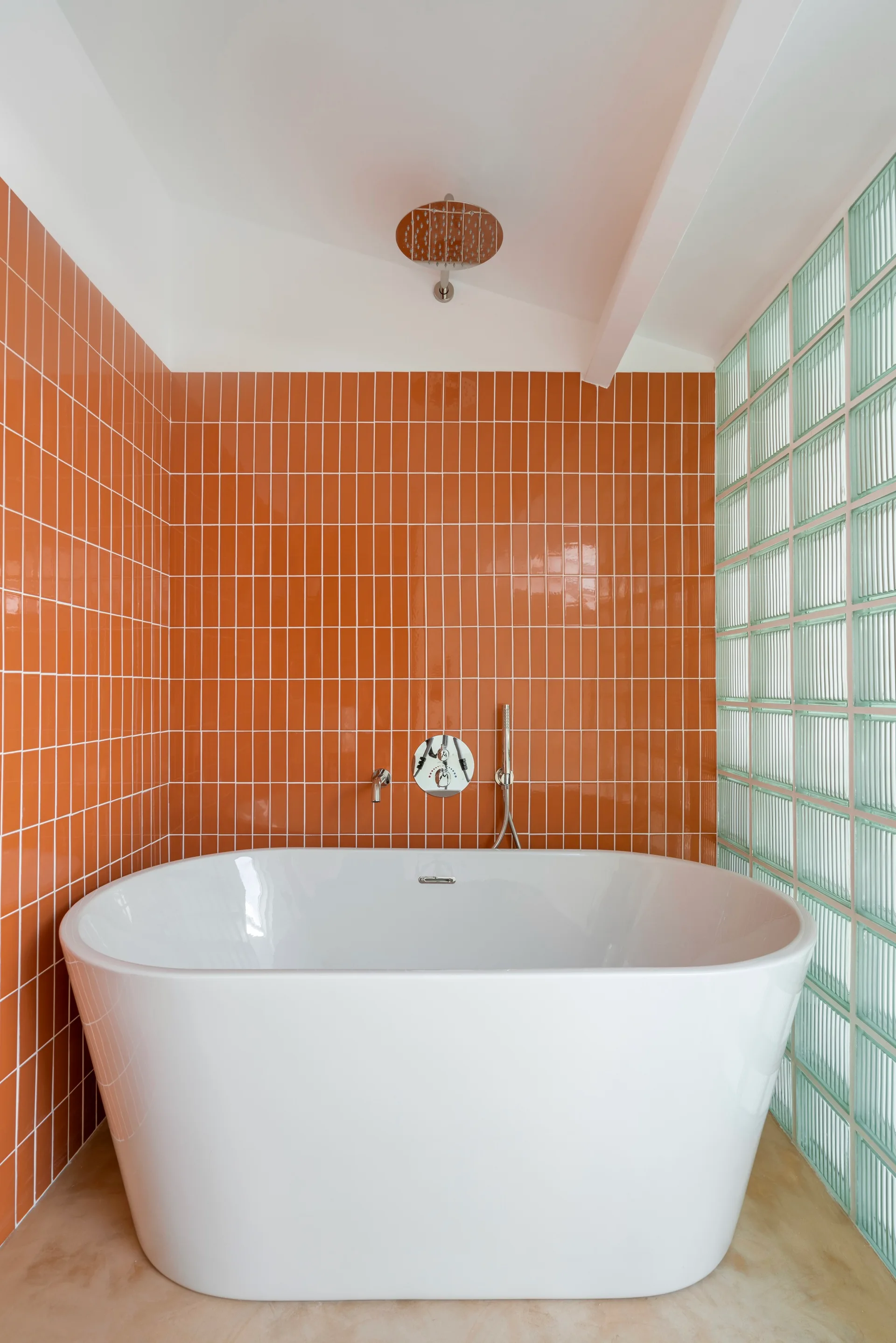 A white freestanding bathtub stands against a glossy orange vertical tile wall next to a translucent glass block partition.