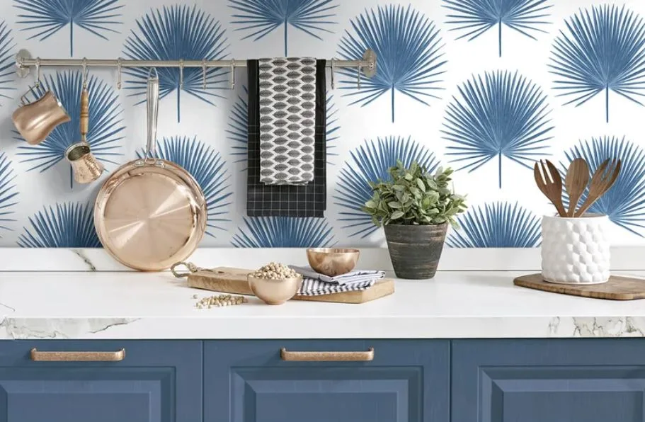 A kitchen counter with blue paneled cabinets, a marble countertop, and blue palm leaf wallpaper with hanging copper pans.