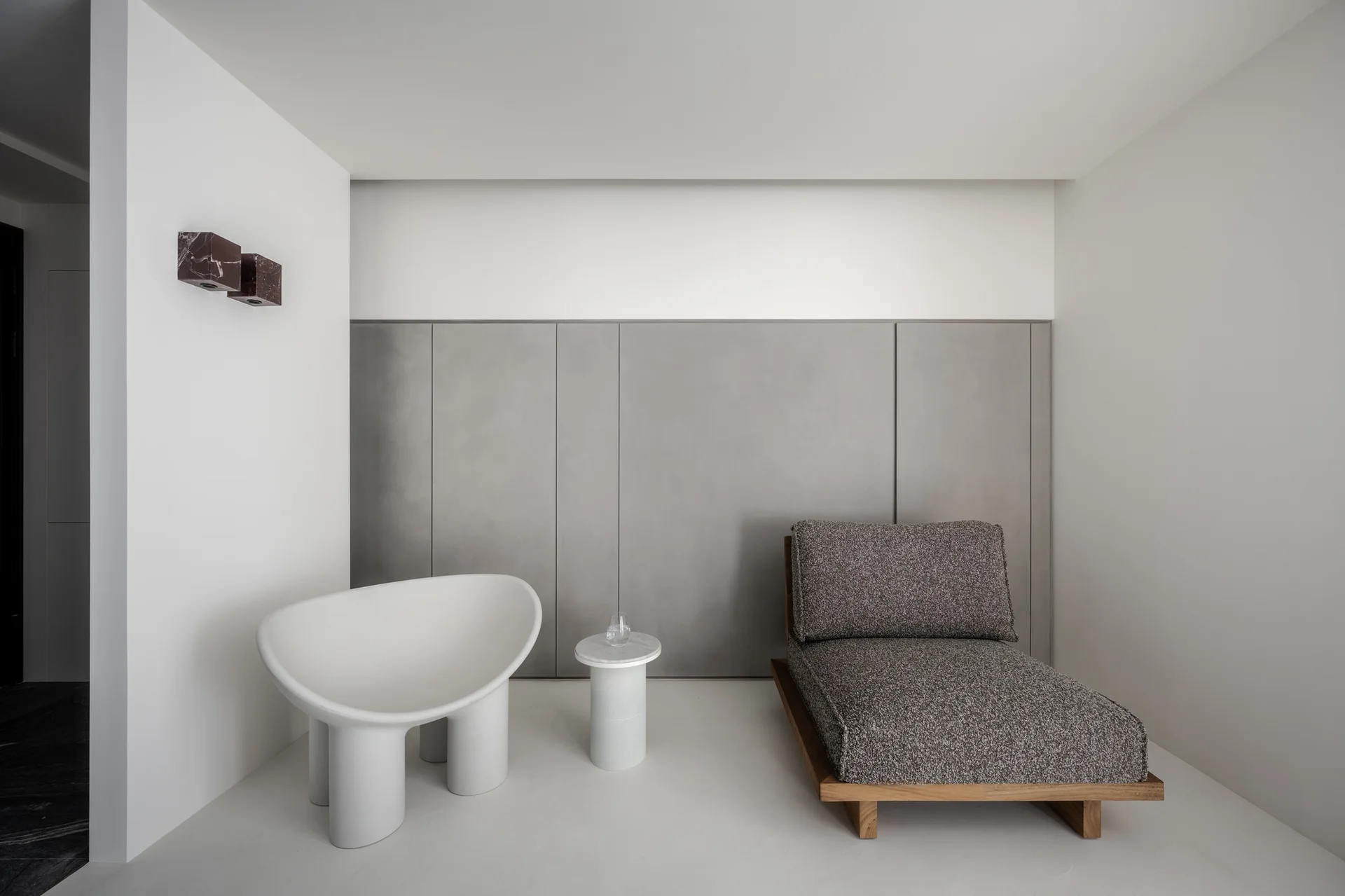 A minimalist seating area features a grey fabric chaise longue, a white sculptural armchair, and a small marble side table against grey wall paneling.