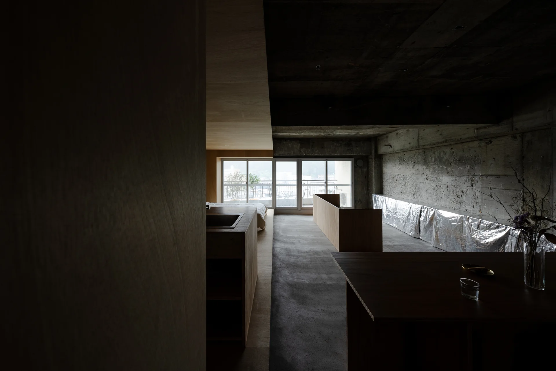 A minimalist industrial apartment features raw concrete walls and ceiling, low wooden partitions, and a large sliding glass door opening to a balcony.