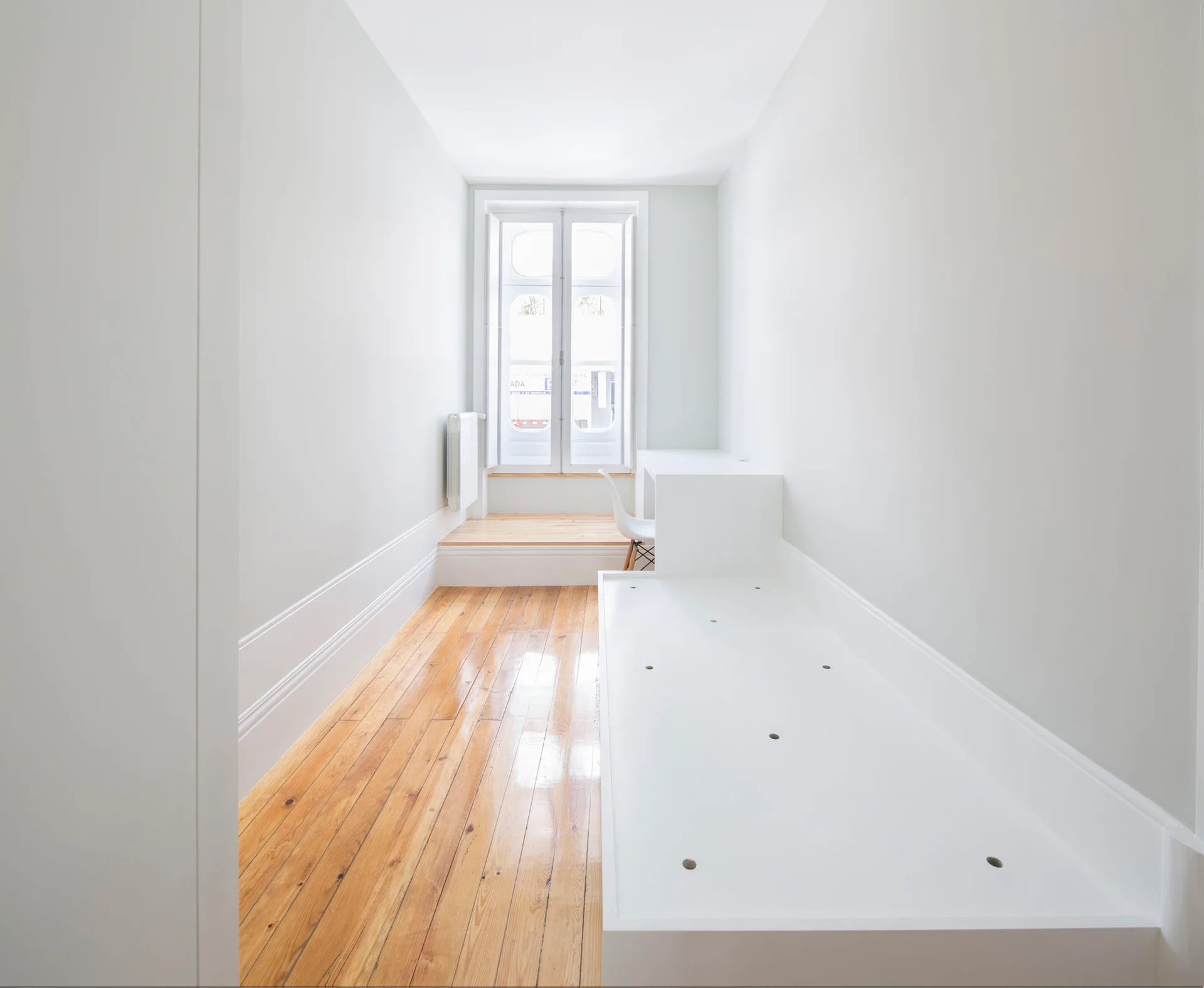 A narrow minimalist bedroom features polished wood floors and a white built-in bed unit that transitions into a study desk by the window.