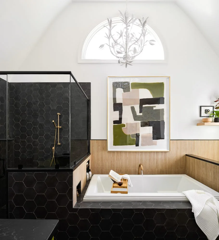 A modern bathroom features a white bathtub surrounded by black hexagonal tiles next to a light wood slat wall under an arched window.