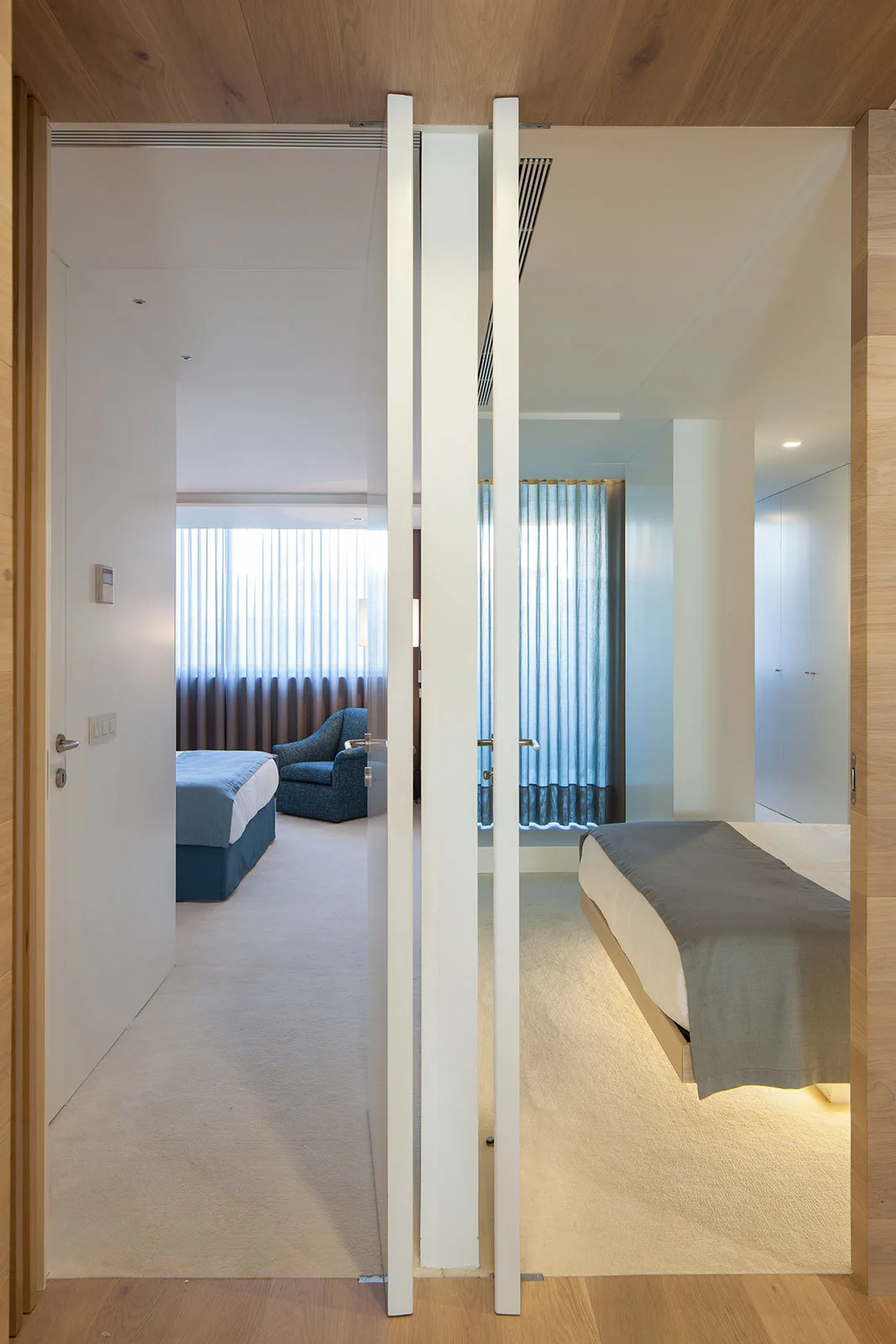 A view through a central doorway showing two minimalist bedrooms with neutral carpets, a wood ceiling, and warm under-bed lighting.