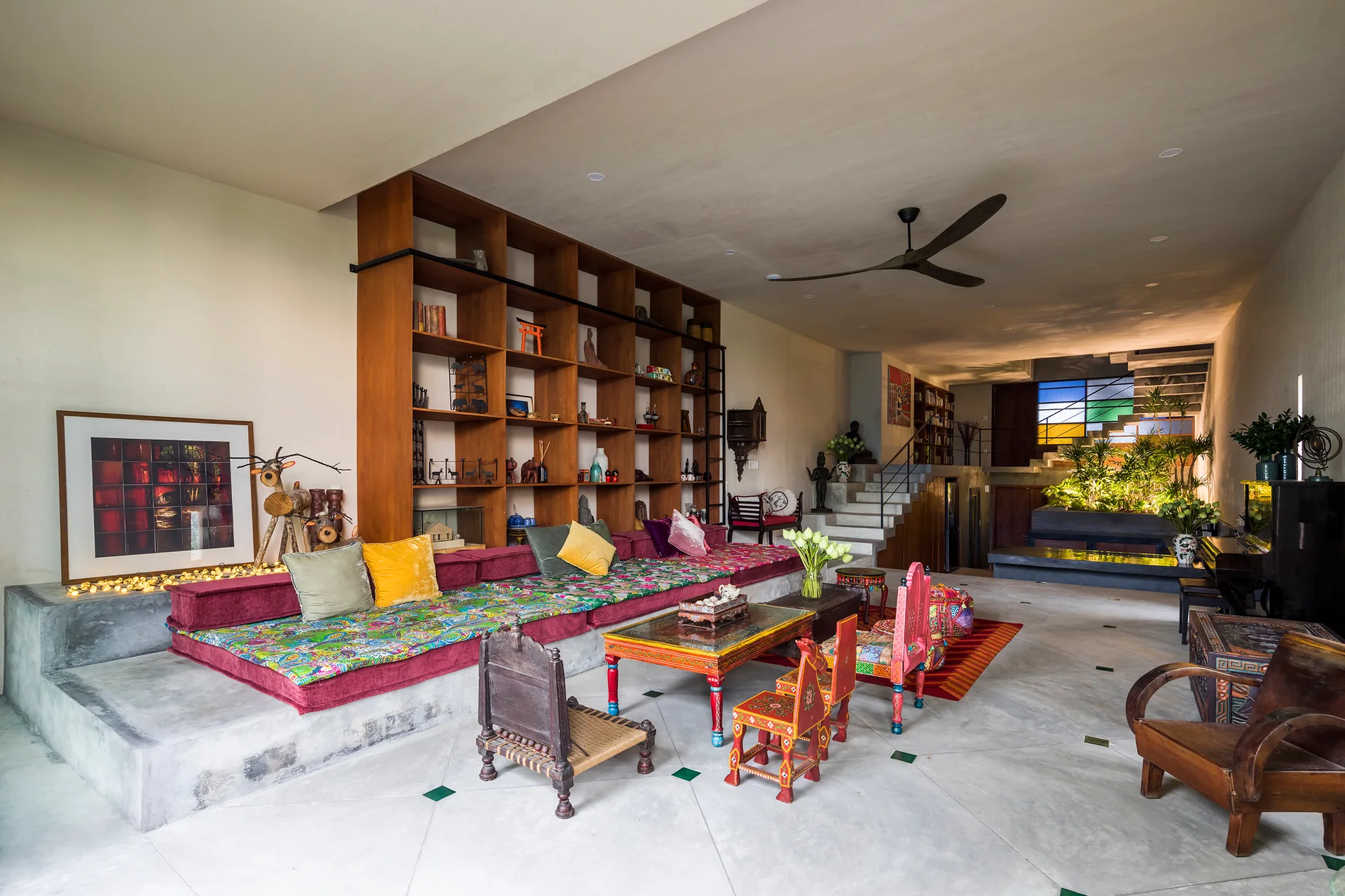 An eclectic living room featuring a raised concrete platform with patterned cushions, a large wooden grid shelving unit, and colorful low chairs.