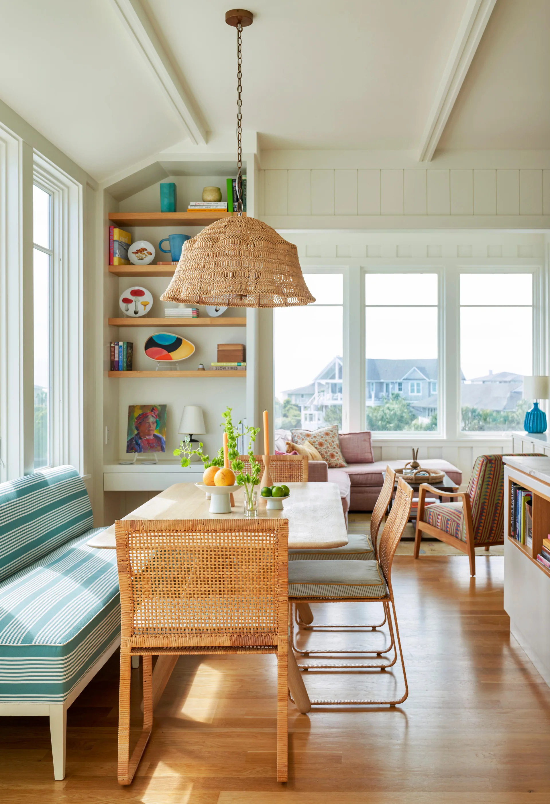 A sunlit dining area features a light wood table with woven wicker chairs, a blue striped upholstered bench, and a large wicker pendant light.