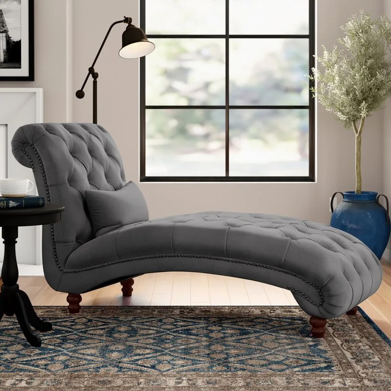 A tufted grey chaise lounge with nailhead trim sits on a patterned blue rug next to a black floor lamp and a blue ceramic planter.