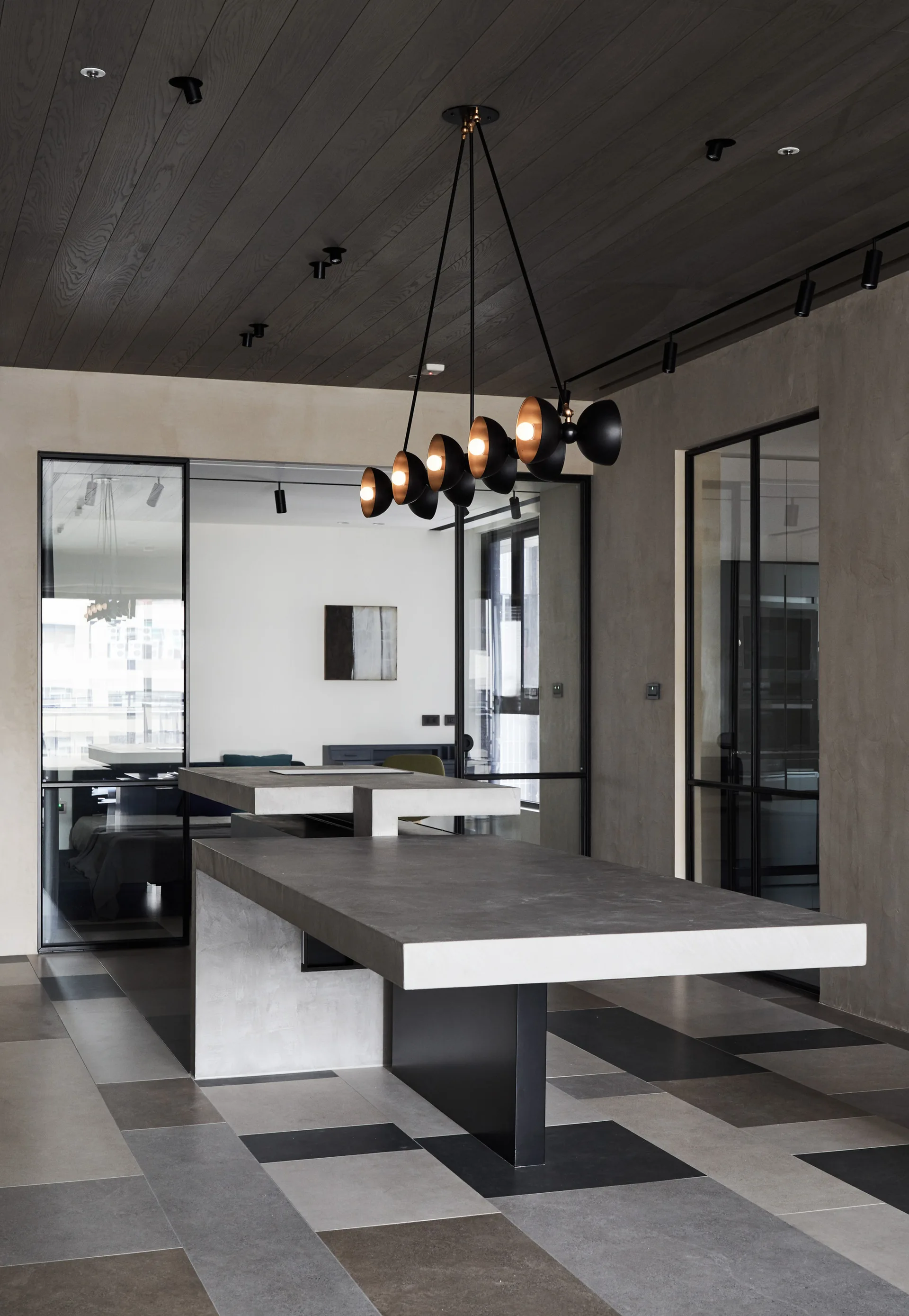A modern kitchen space features a multi-level grey concrete island beneath a black sculptural pendant light, with black metal-framed glass partitions.