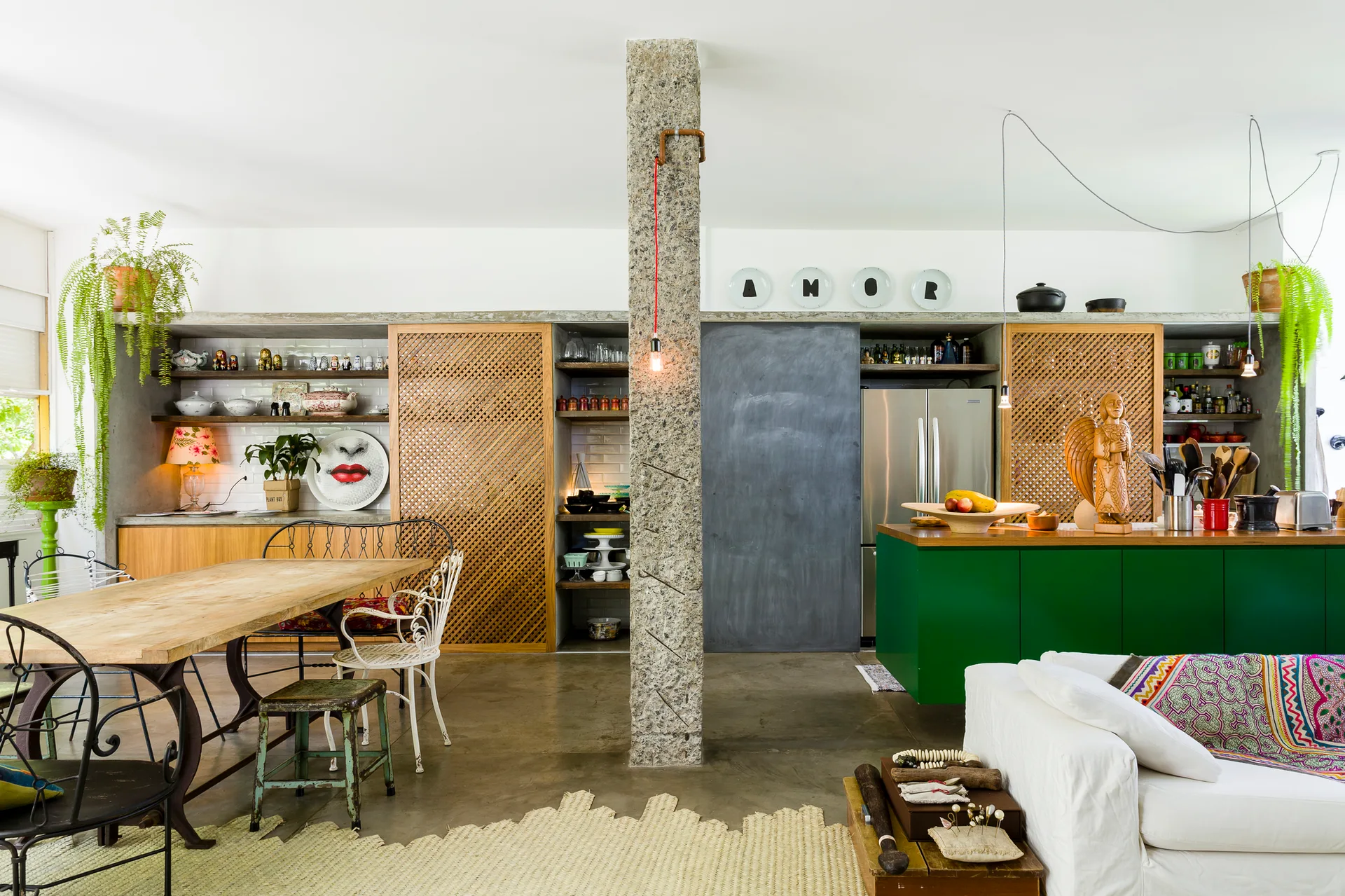 An open kitchen featuring a green island, wood lattice cabinetry, a rustic dining table, and a prominent rough stone support column.
