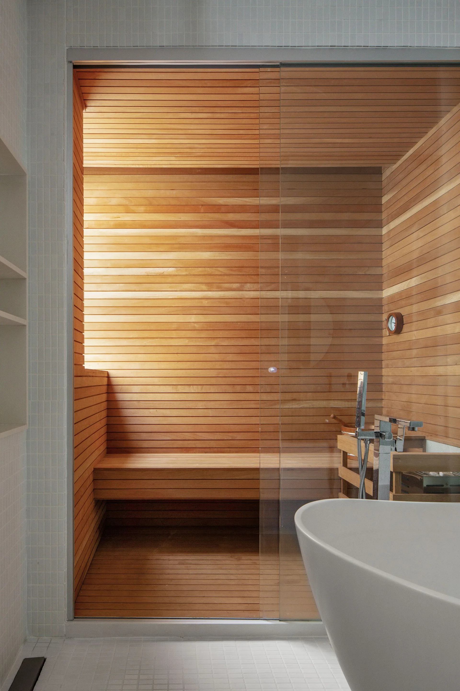 A modern home sauna with horizontal wood slats and a glass partition wall next to a white freestanding bathtub on tiled flooring.