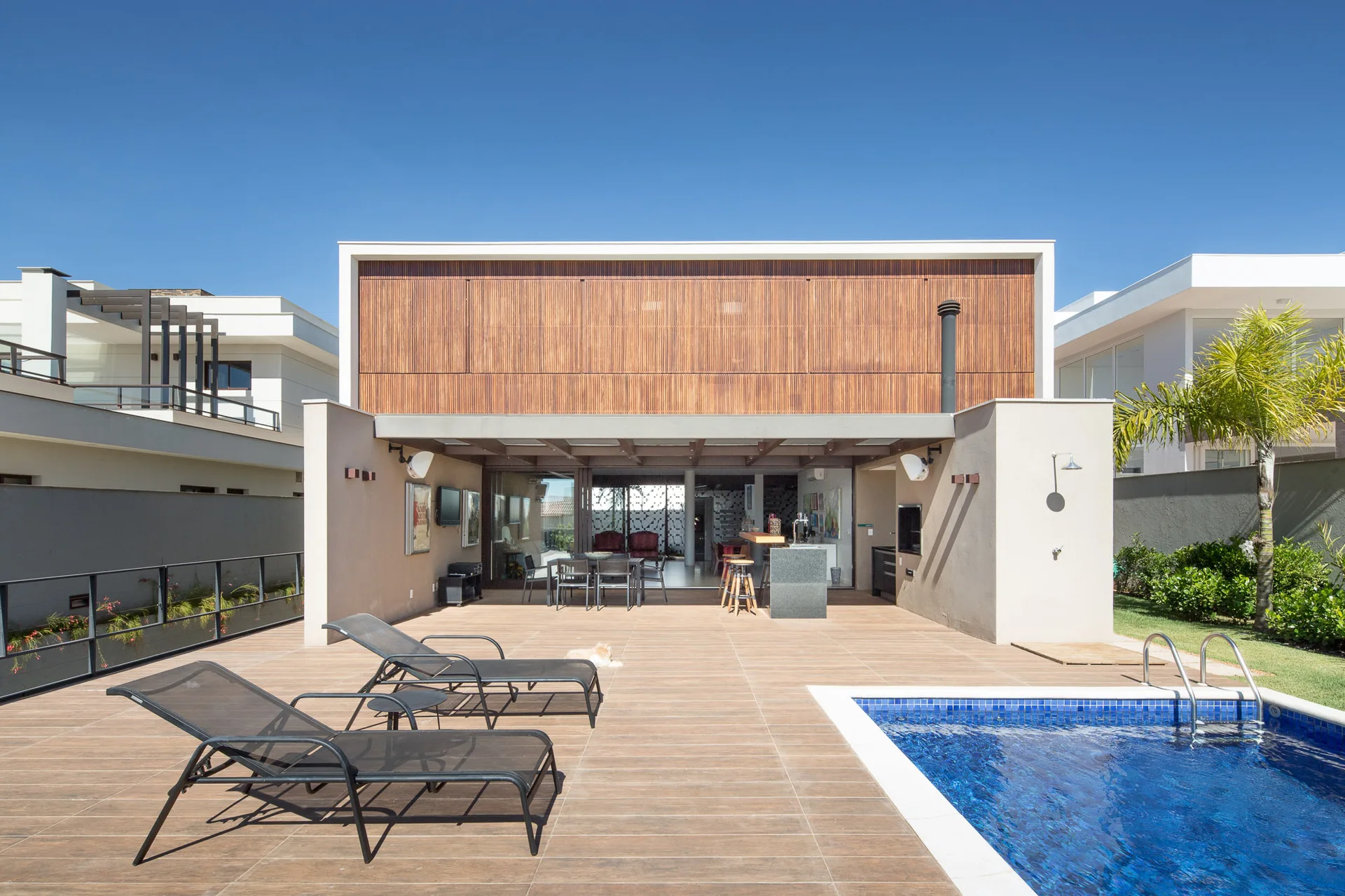 A modern outdoor patio features a blue swimming pool, wood-look tiled deck with lounge chairs, and an outdoor kitchen under a wood-slat facade.