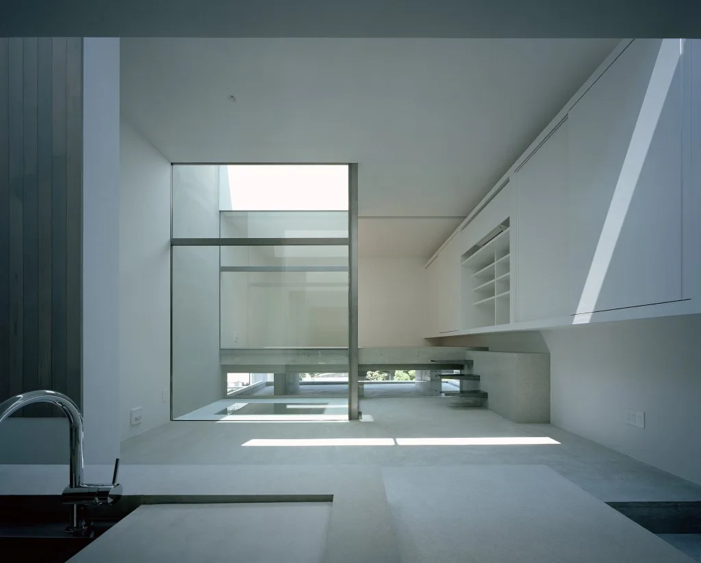 A minimalist white kitchen features handleless cabinets, a concrete floor with direct sunlight, and a large structural glass partition with stairs.
