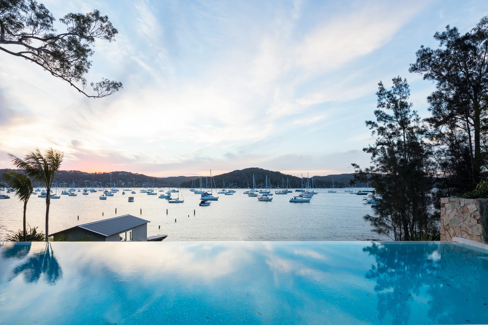 An infinity edge swimming pool with blue tiles overlooks a bay filled with anchored sailboats at dusk.