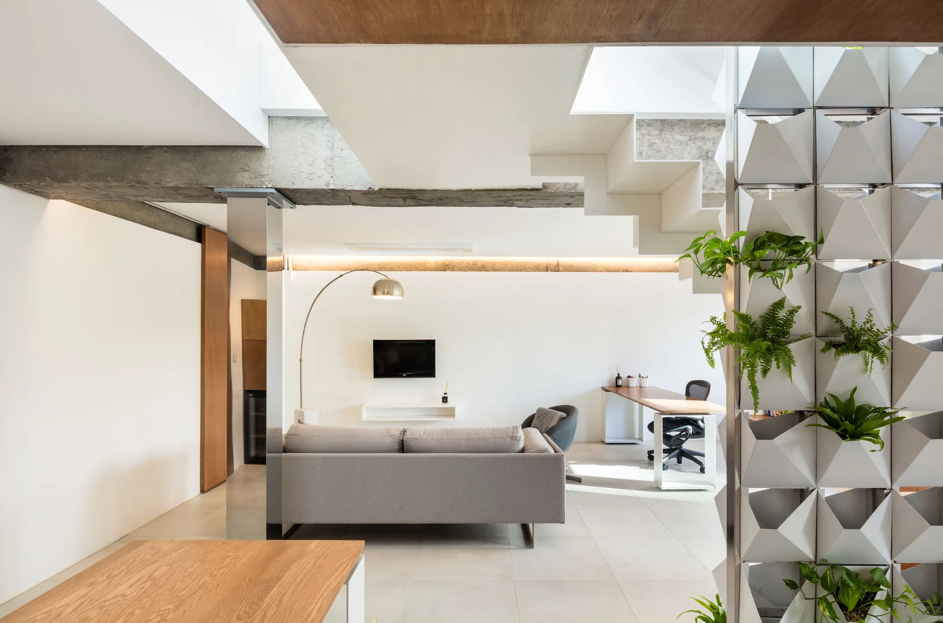 A modern open-plan living room and home office featuring a gray sofa, a wood desk, an exposed concrete beam, and a white geometric planter wall.
